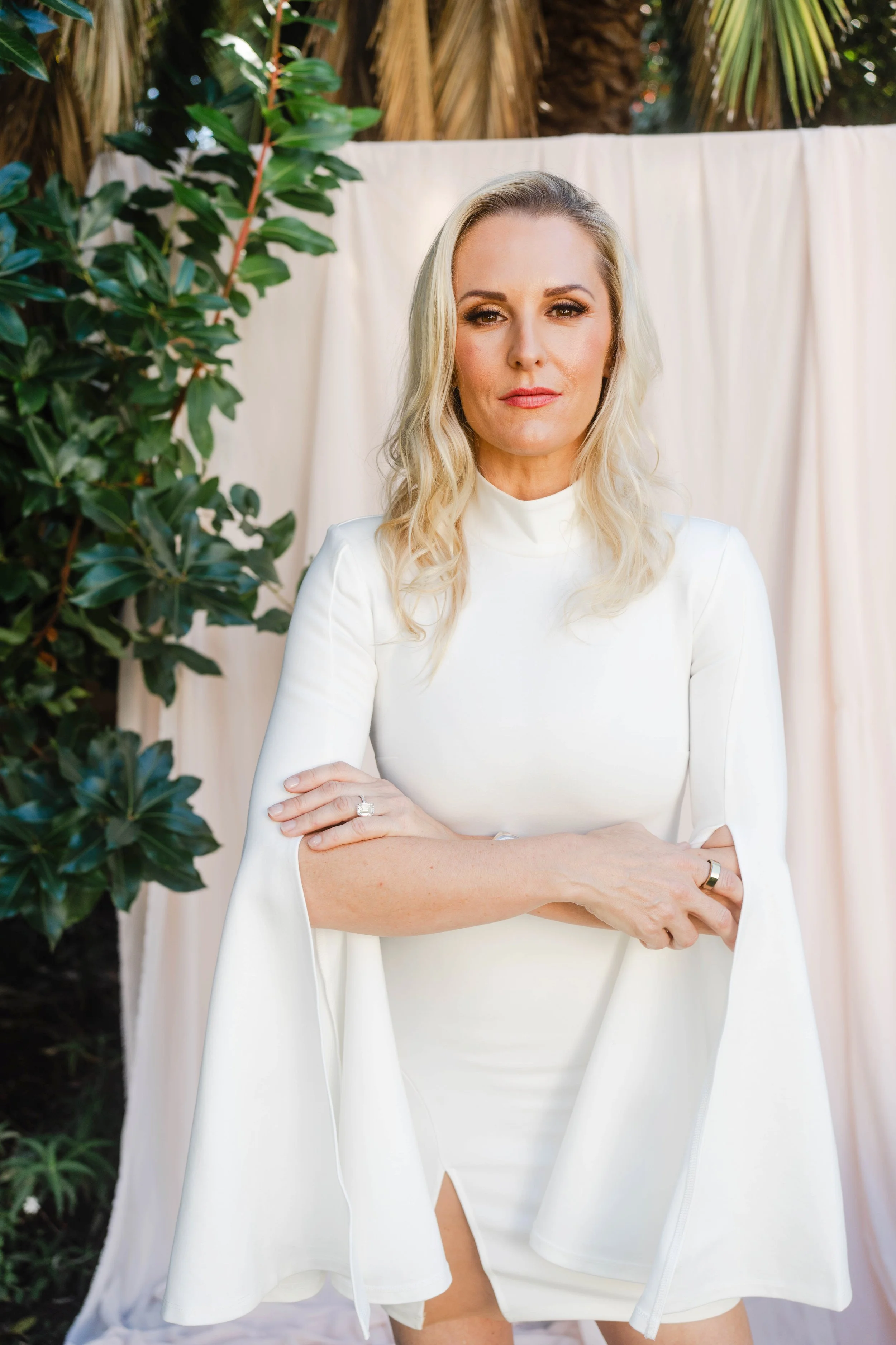 Woman with blonde hair in a white dress with cape standing outdoors in front of a white backdrop and green foliage.