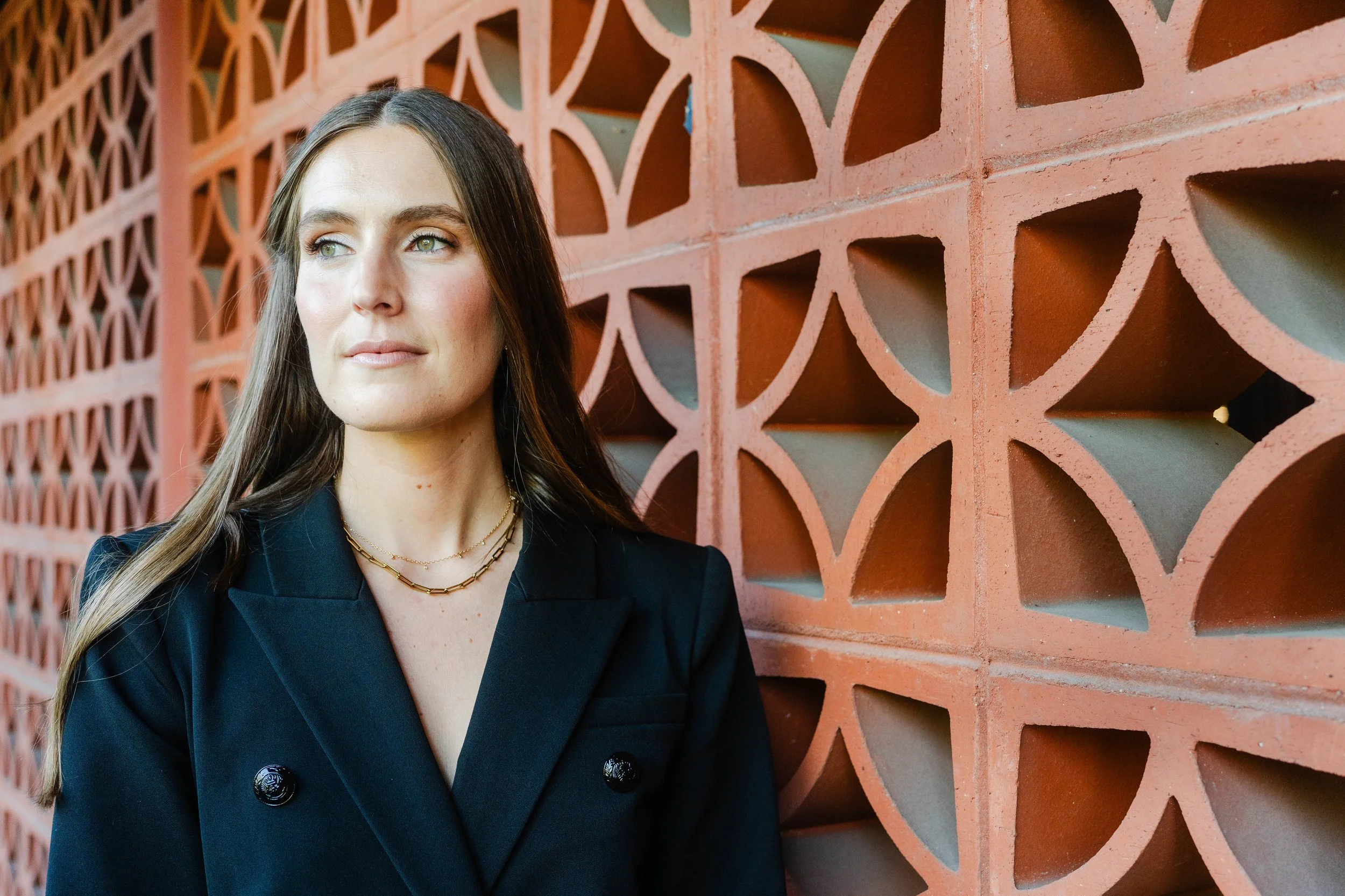 A woman with long brown hair wearing a black blazer and gold necklaces stands in front of an ornate, geometric-patterned brick wall.