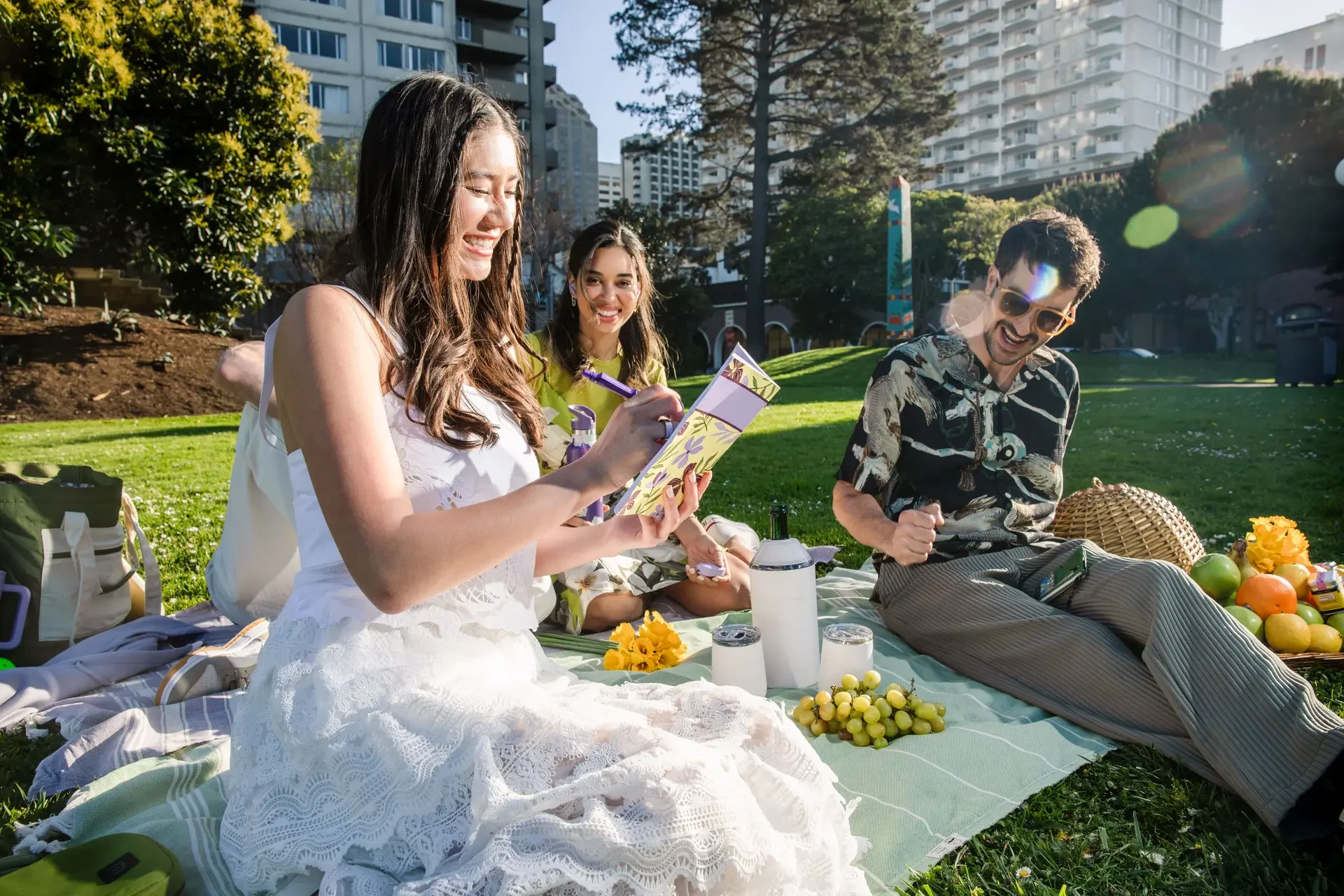 Three friends having a picnic on a blanket in a park surrounded by tall buildings, with one woman writing on a notebook and two people smiling and laughing.