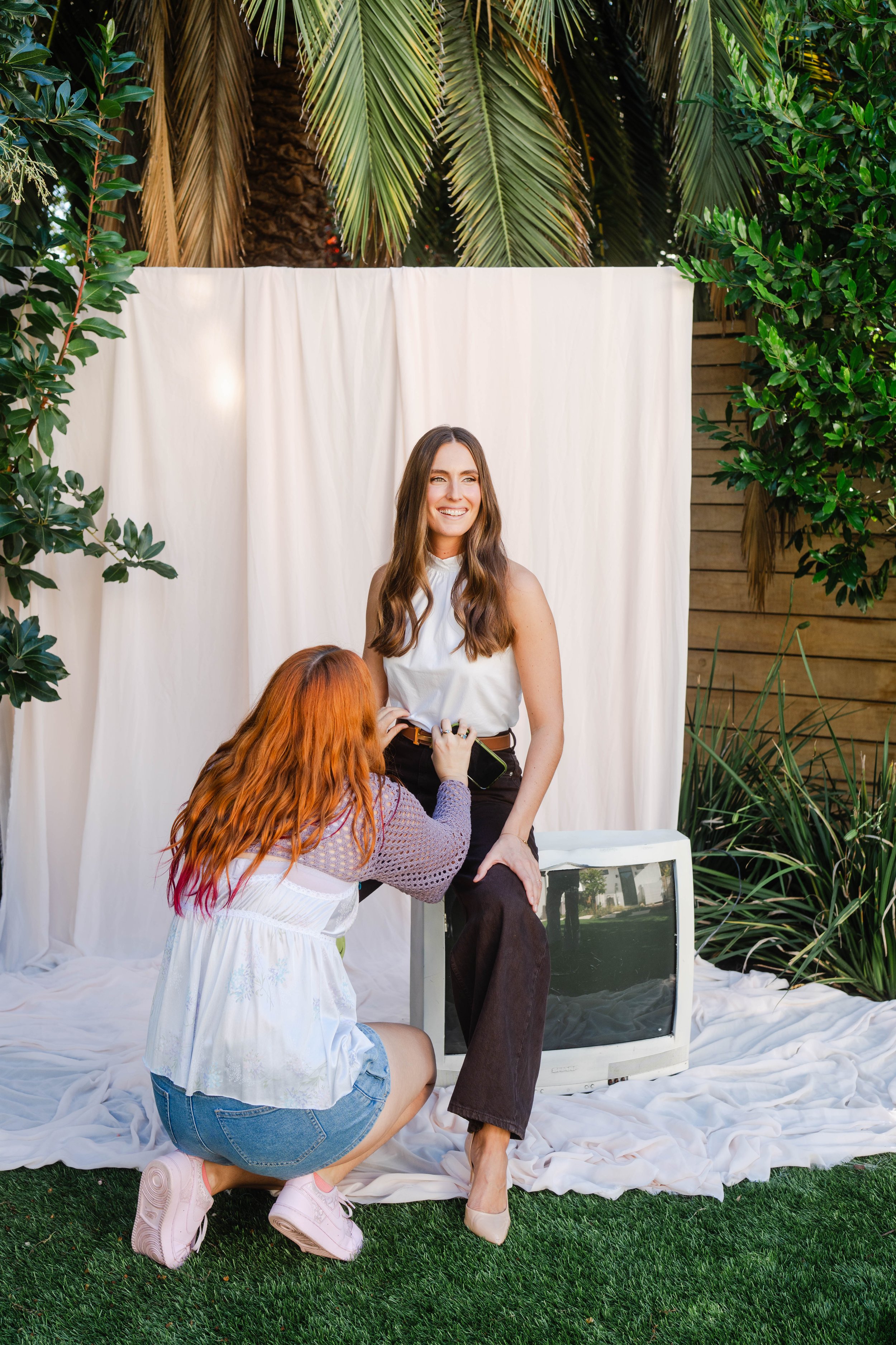 A woman with long brown hair sitting on a white chair, getting a portrait taken outdoors. A photographer with red hair and pink shoes is kneeling in front, taking her picture. The scene is against a white fabric backdrop surrounded by greenery, including palm fronds and bushes.