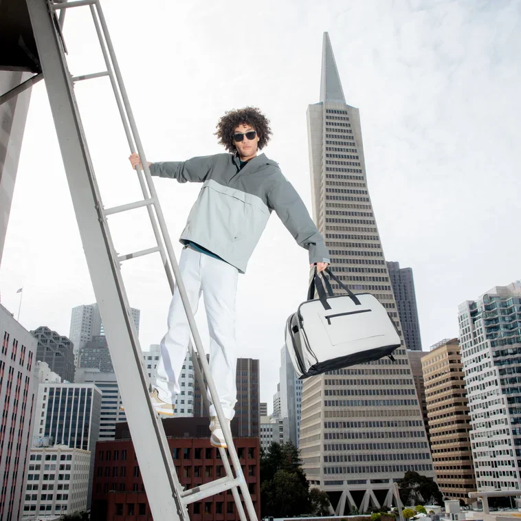 A person standing on a ladder in a cityscape holding a white duffle bag, with the Transamerica Pyramid in San Francisco in the background.