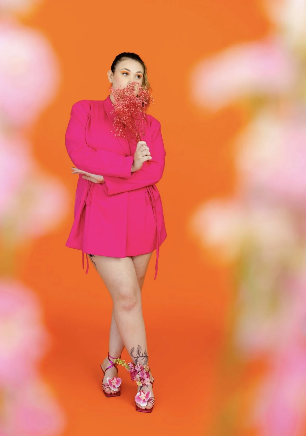 A woman in a hot pink dress holding a bouquet of pink flowers against an orange background with pink and white floral decorations in the foreground.