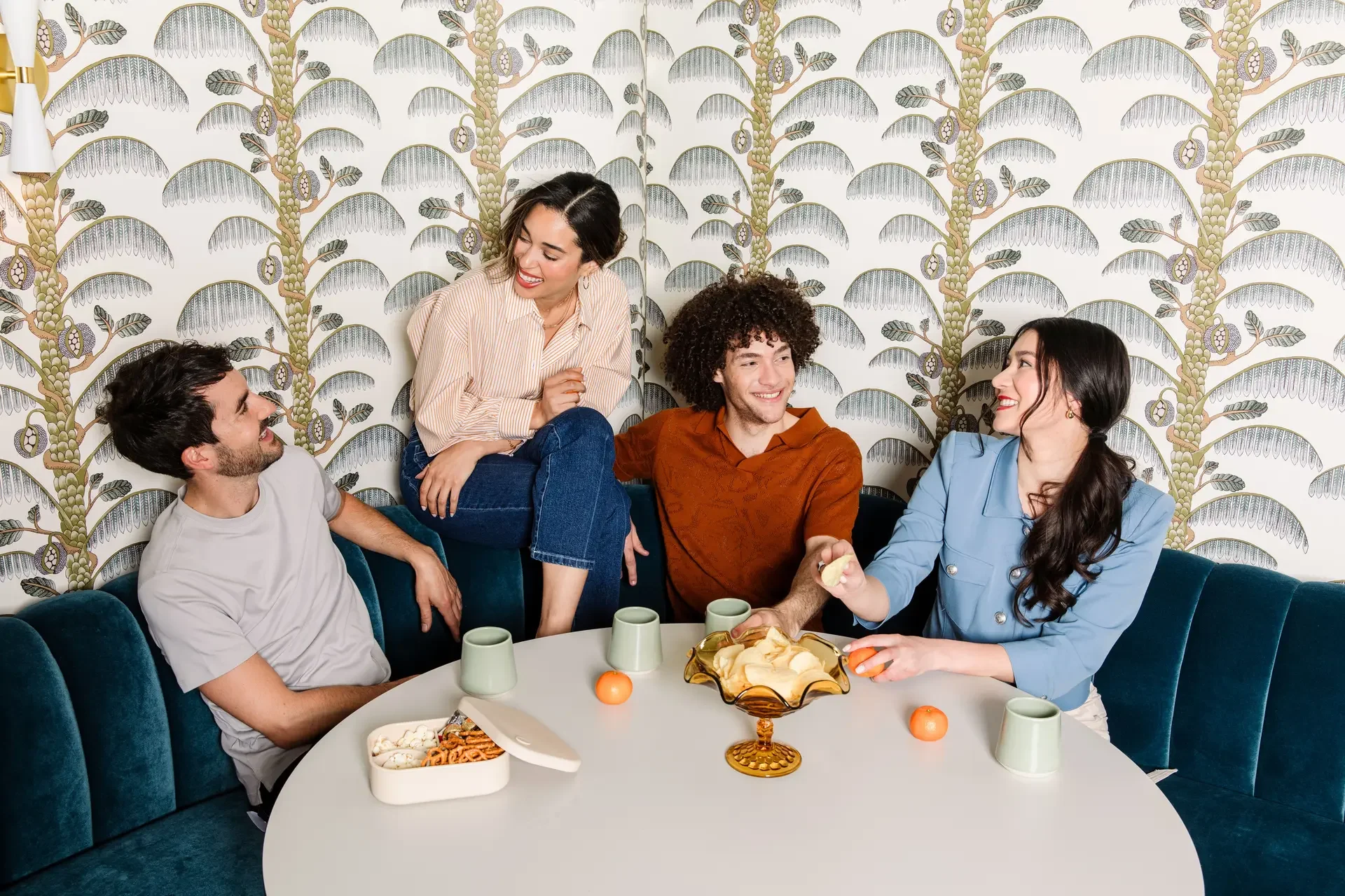 Group of five friends enjoying a casual gathering around a table, chatting and smiling, in a living room with patterned wallpaper.