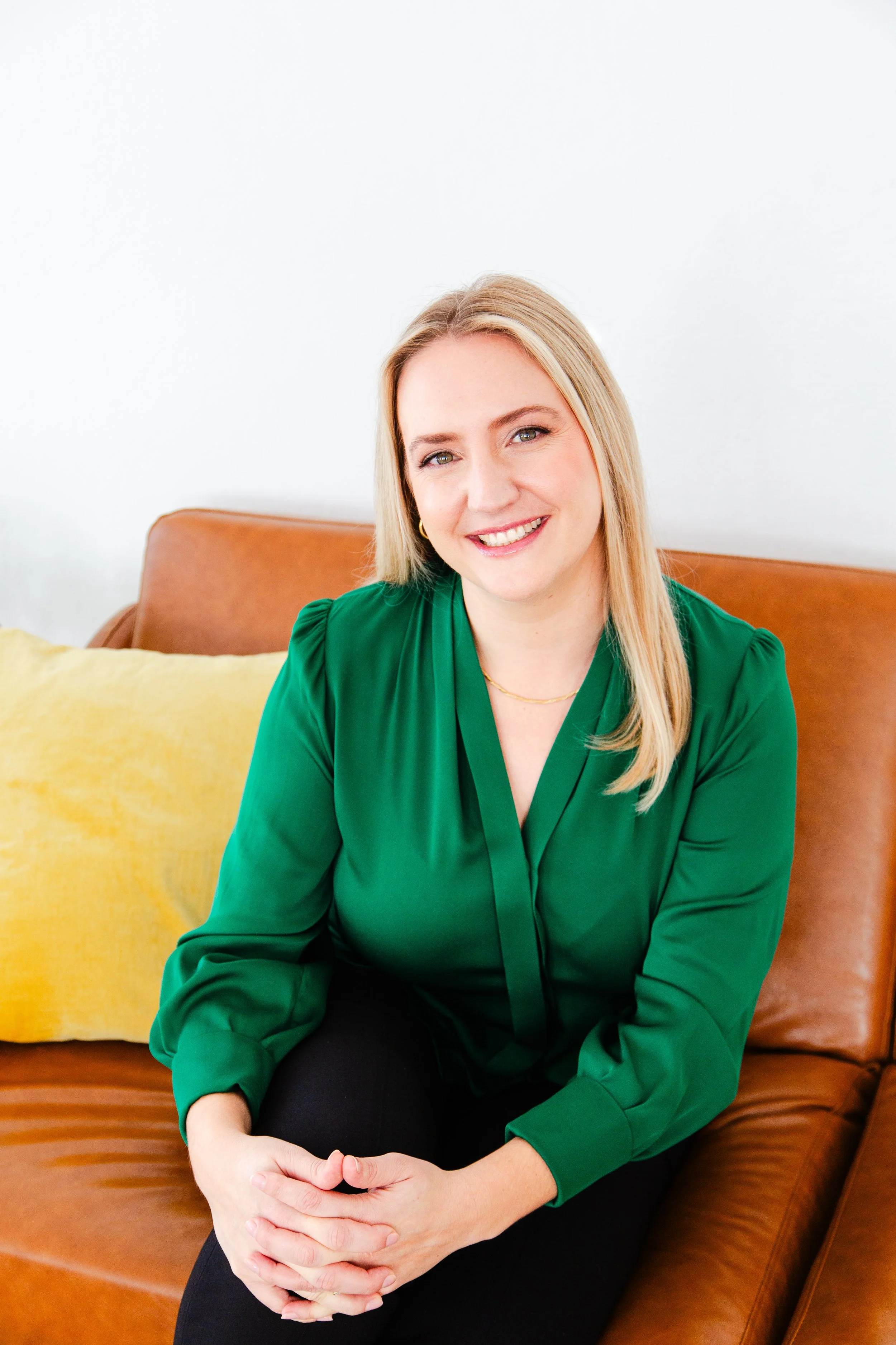 A smiling woman with blonde hair, wearing a green blouse, sitting on a brown leather sofa with a yellow pillow, in front of a white wall.