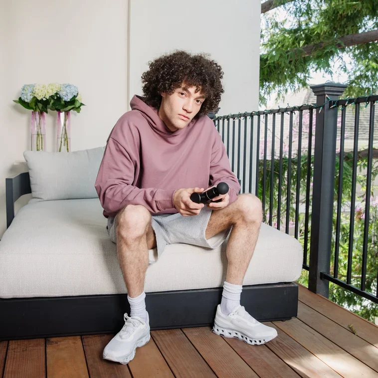 A young man with curly hair sitting on a porch swing, holding a microphone, wearing a mauve hoodie, gray shorts, white socks, and sneakers, with a railing and trees in the background.