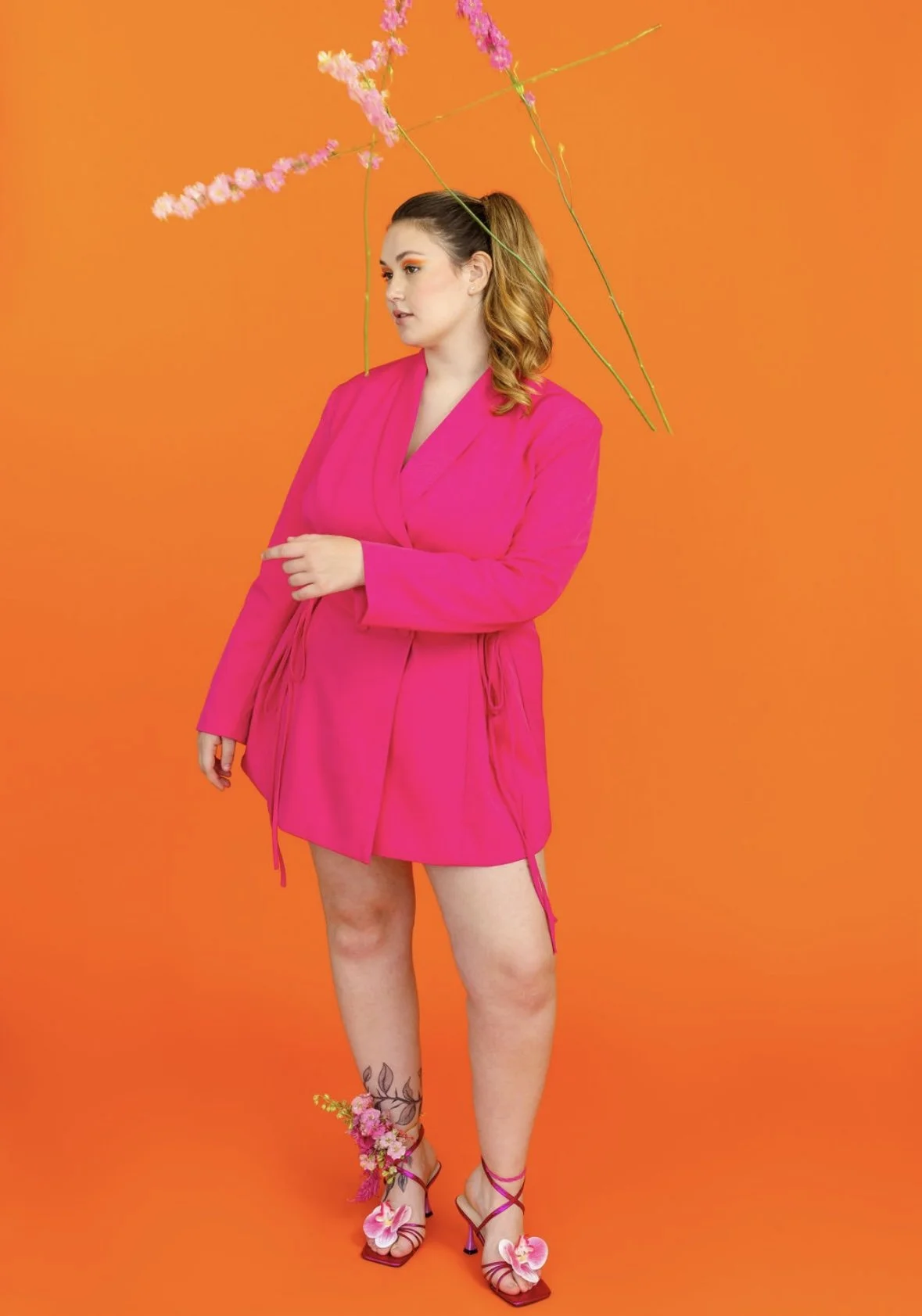 A woman standing against an orange background, dressed in a bright pink blazer and skirt, holding a stick with pink flowers over her head, with floral decorations on her ankle and pink high heels with flowers.