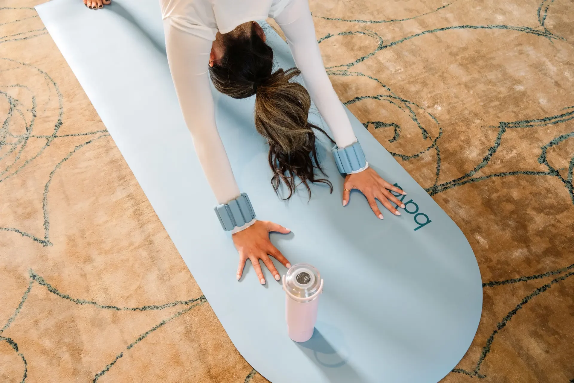 A woman practicing yoga on a blue mat, wearing light-colored clothing, sunglasses on her head, and multi-colored wristbands, with a pink water bottle nearby.