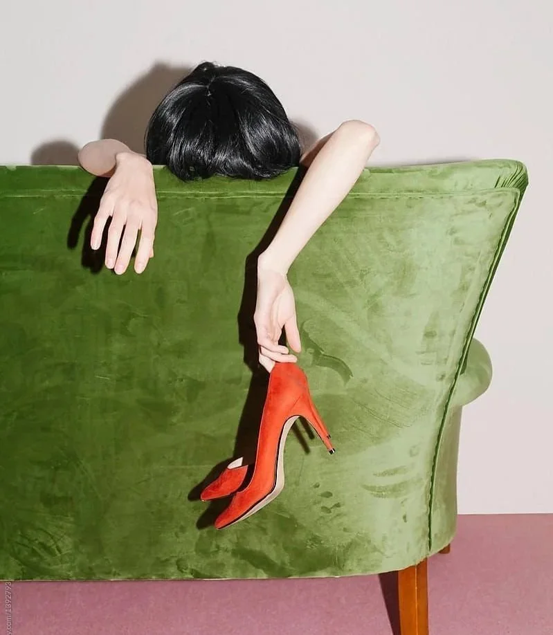 Person hiding behind a green velvet armchair, holding a red high-heeled shoe.