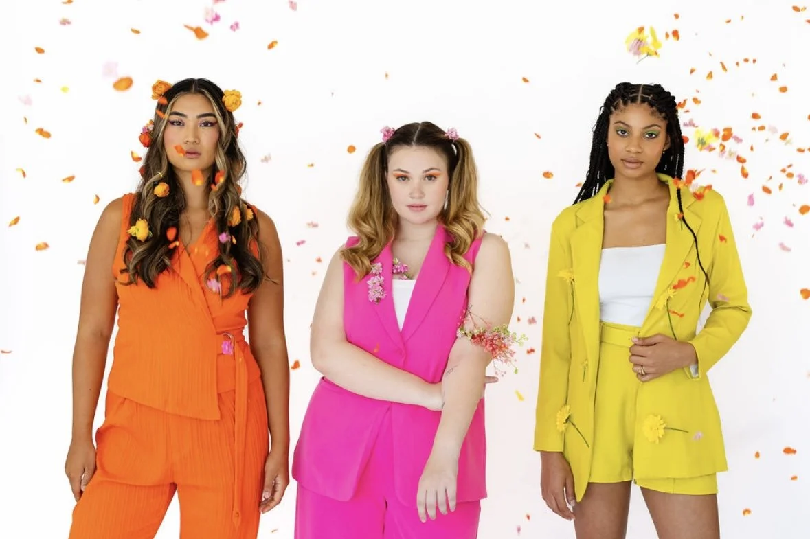 Three women standing together with colorful confetti falling around them, each wearing bright-colored outfits: orange, pink, and yellow.