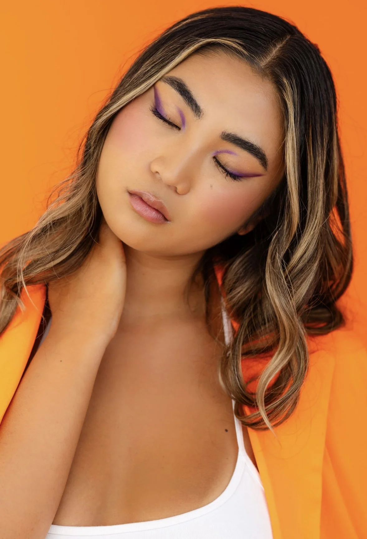 A woman with long wavy brown hair and makeup with purple eyeliner, closed eyes, and a neutral expression, wearing a white top and orange jacket, against an orange background.