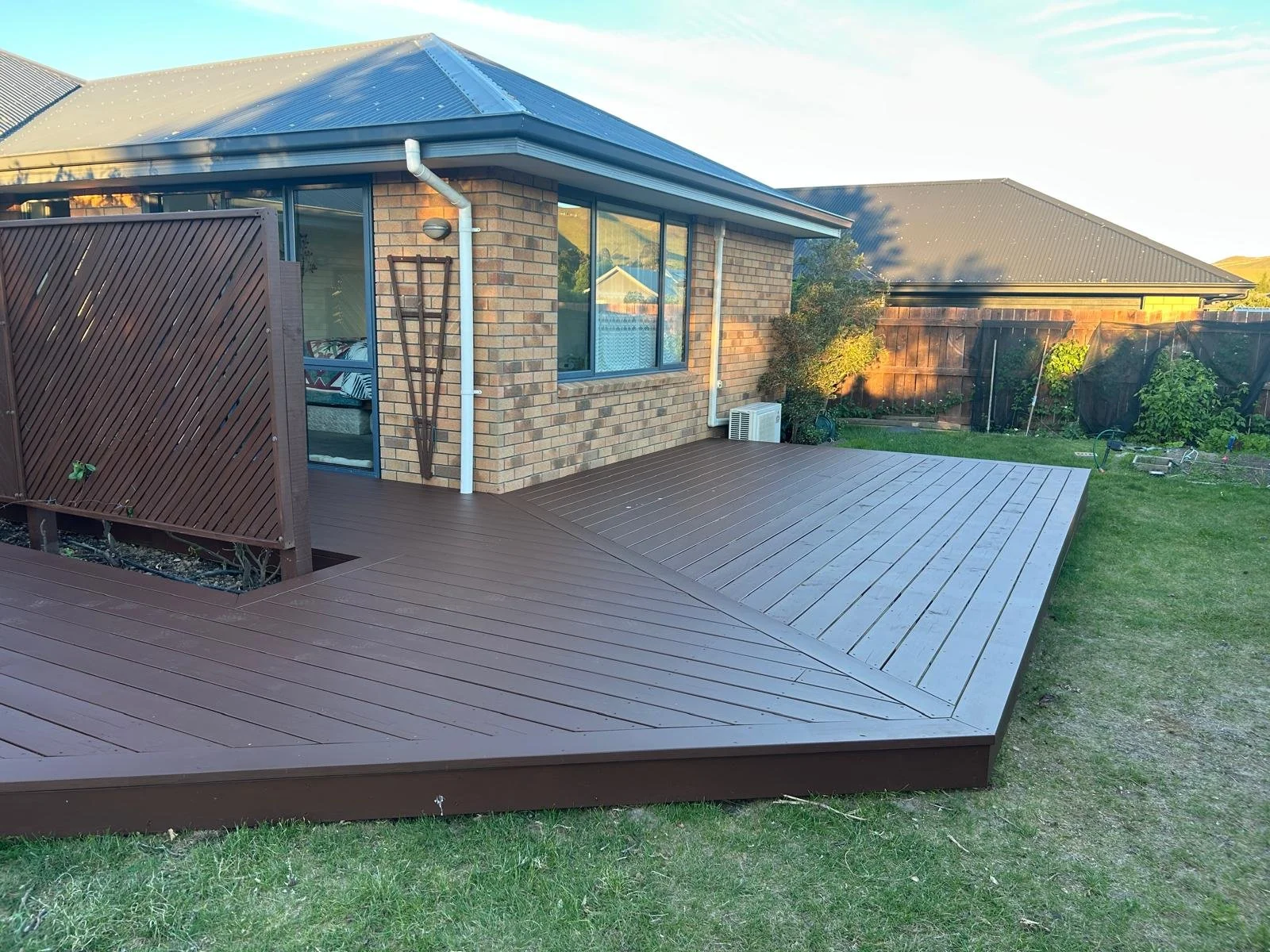 Backyard with a wooden deck attached to a brick house, wooden privacy fence, green grass, and garden plants.