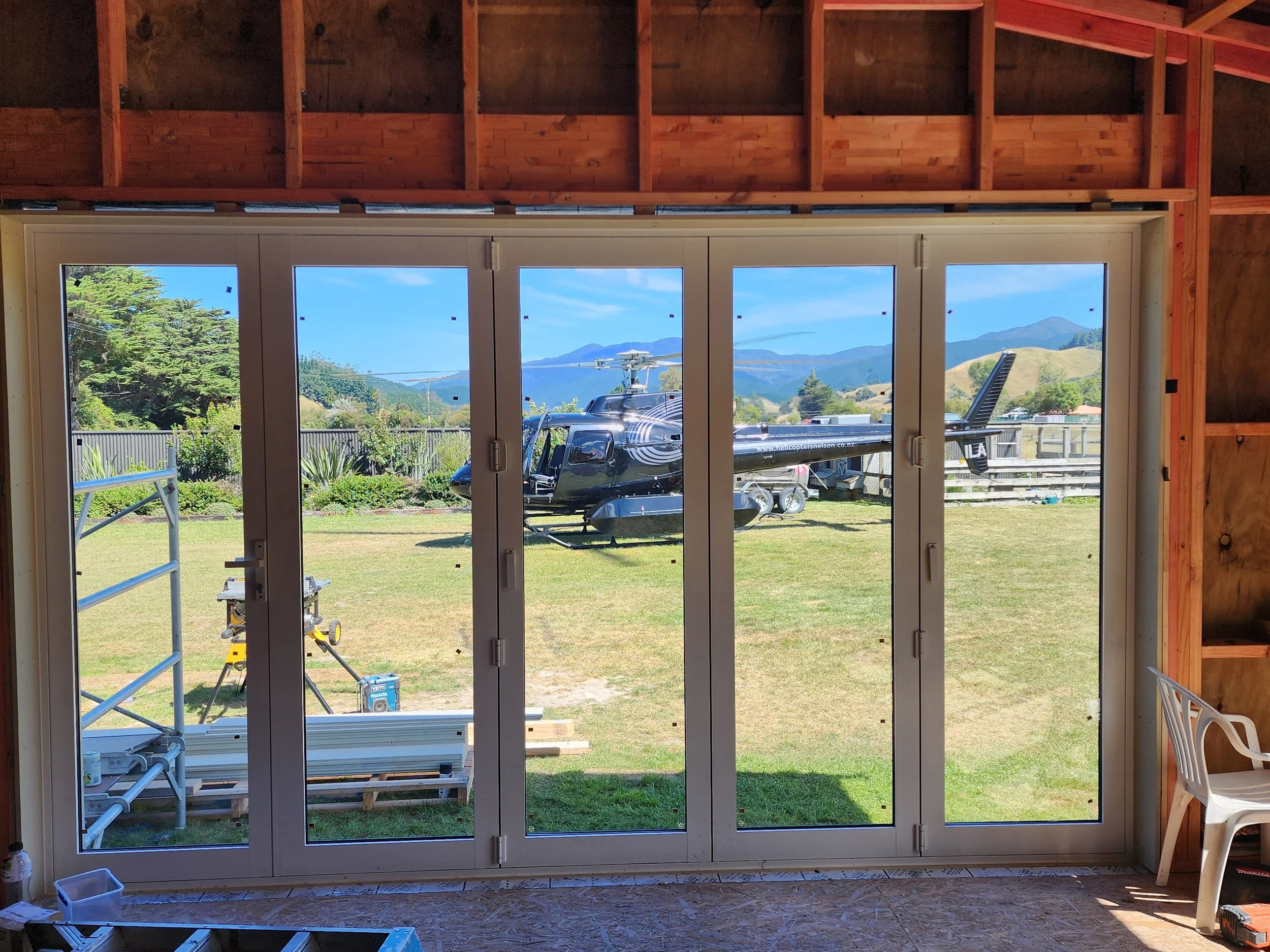 View through glass doors showing a construction or renovation site with tools and equipment inside, and a scenic outdoor landscape with mountains, trees, a helicopter, and a clear blue sky outside.