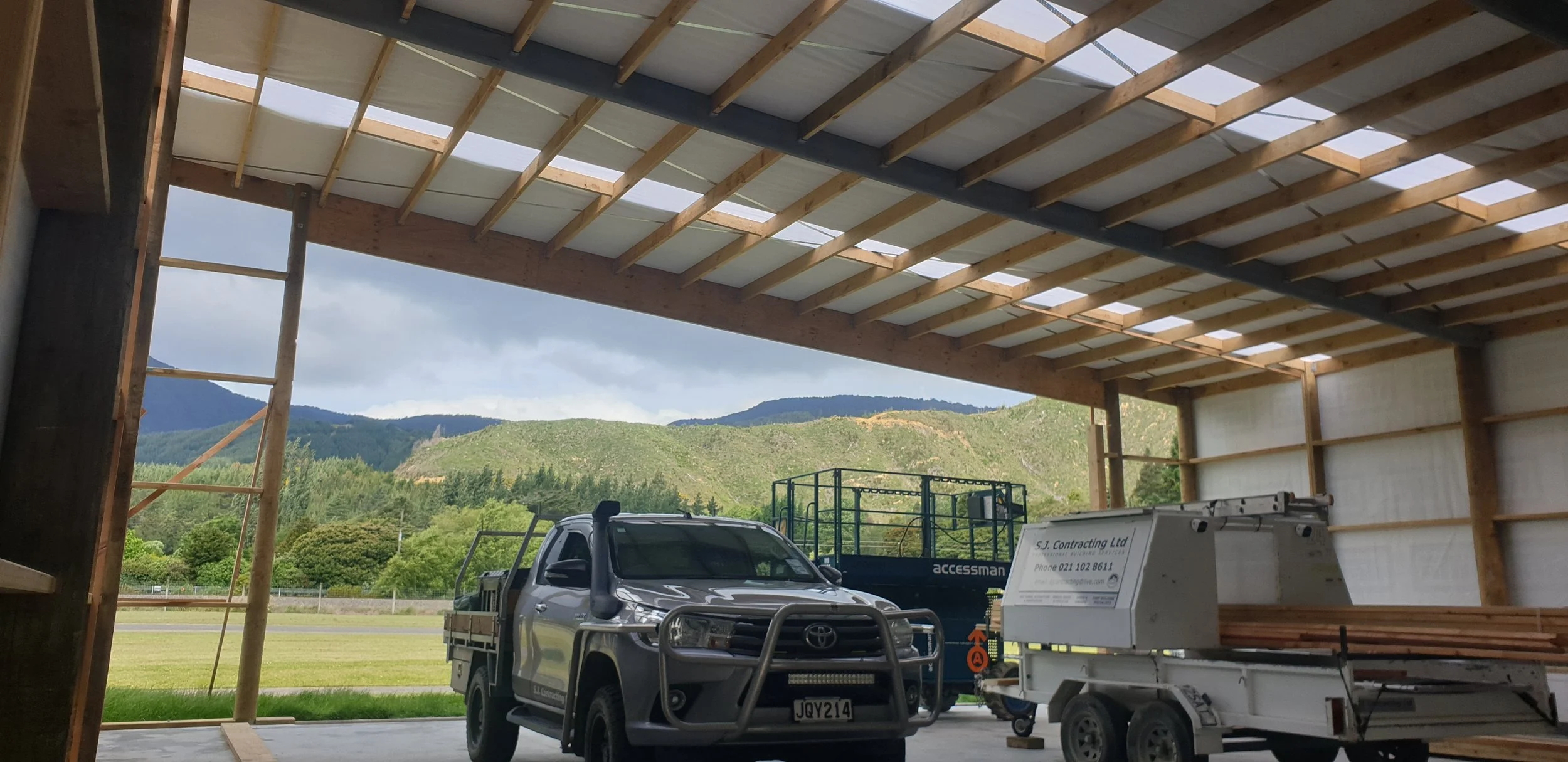 Construction site with an unfinished building frame, trucks, and tools, overlooking green hills and mountains in the distance.