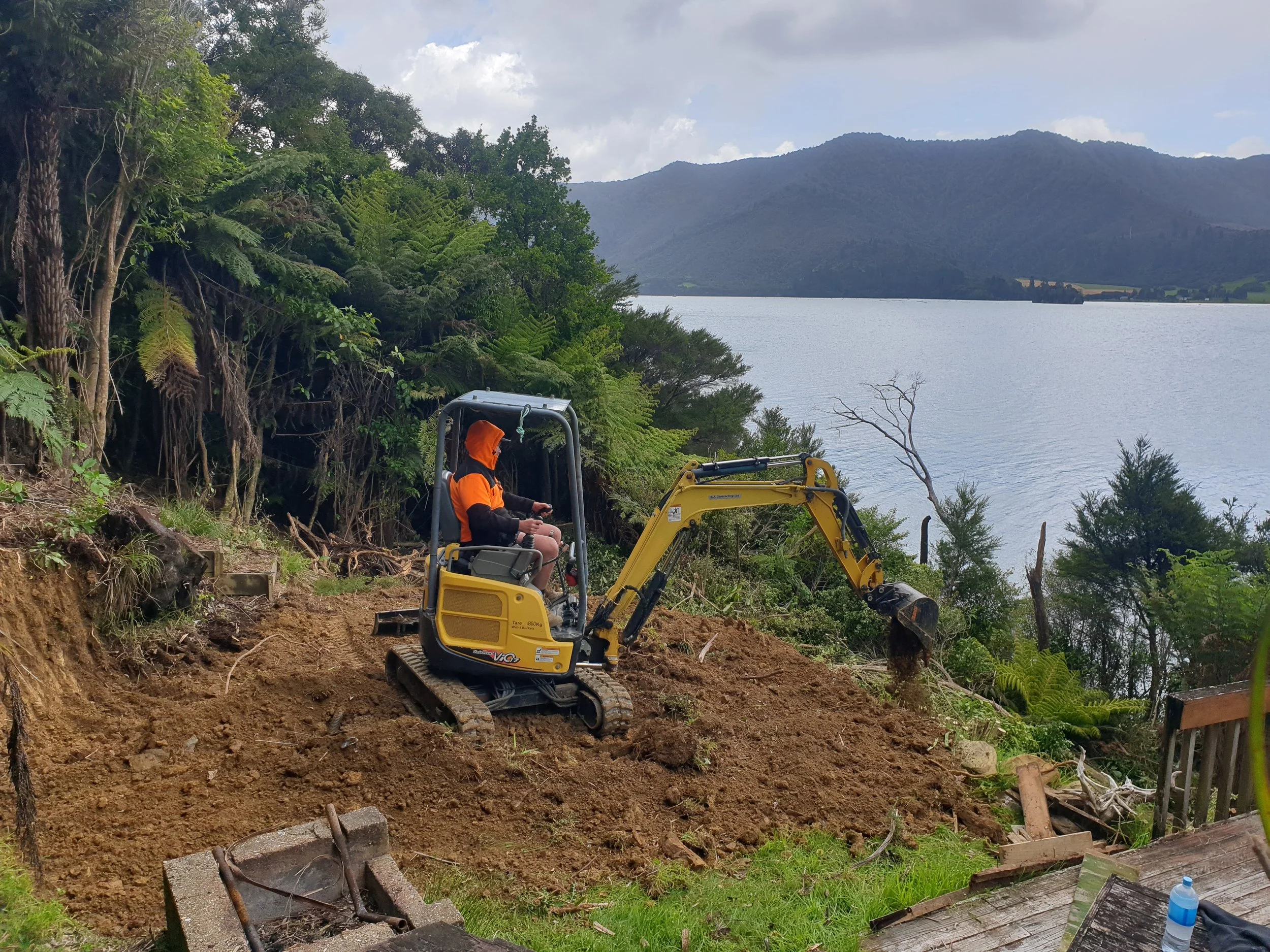 A person in an orange hoodie operating a small yellow excavator on a sloped area of dirt and soil, with lush green trees and a lake in the background.