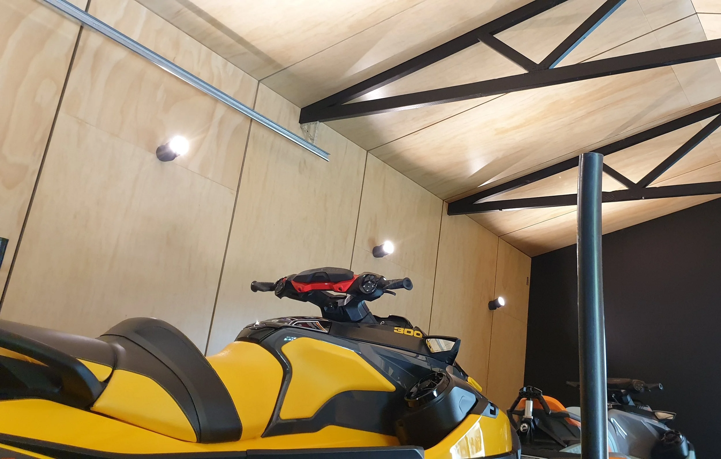 Garage with yellow motorcycle, black motorcycle, and a red and black motorcycle, with wooden and black walls, ceiling lights, and a metal pole.