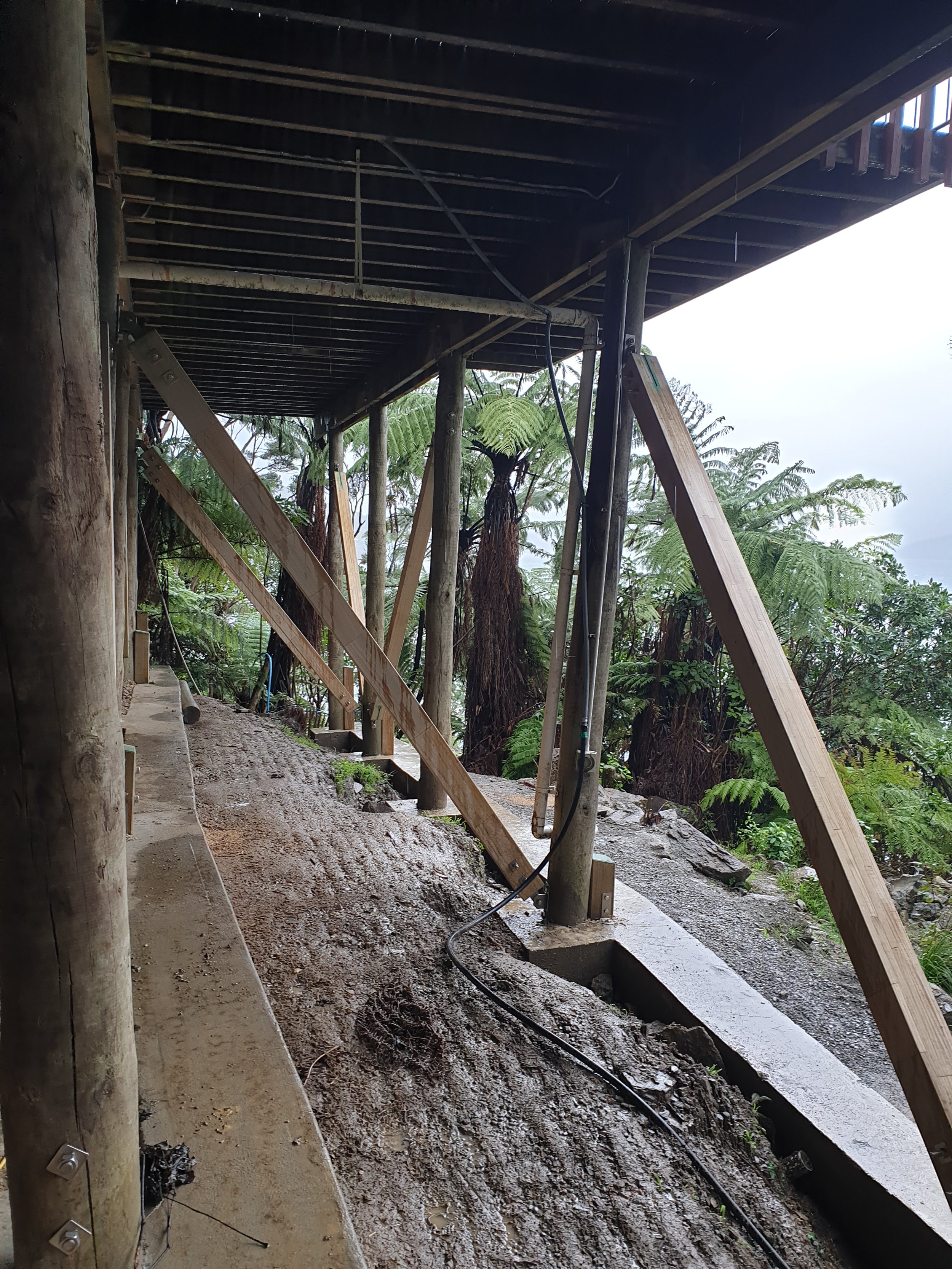 Construction site with wooden supports and a metal structure under a deck, surrounded by lush green trees.