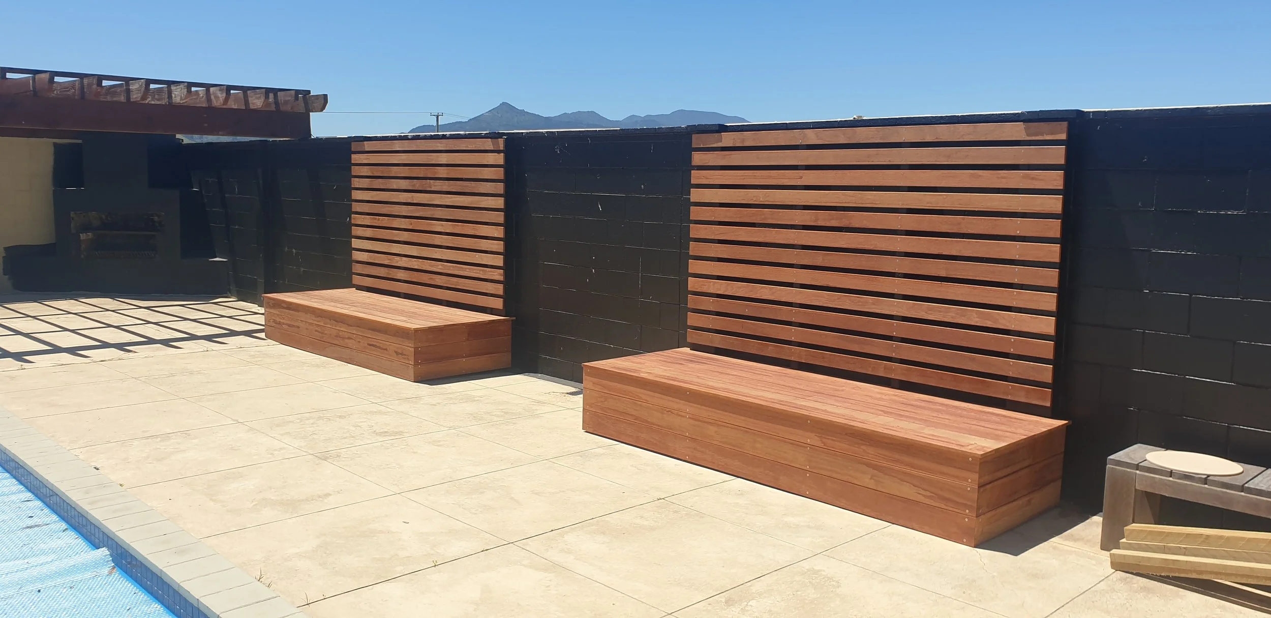 Outdoor patio area with two wooden benches and a wooden privacy screen attached to a black wall, with mountains in the background.