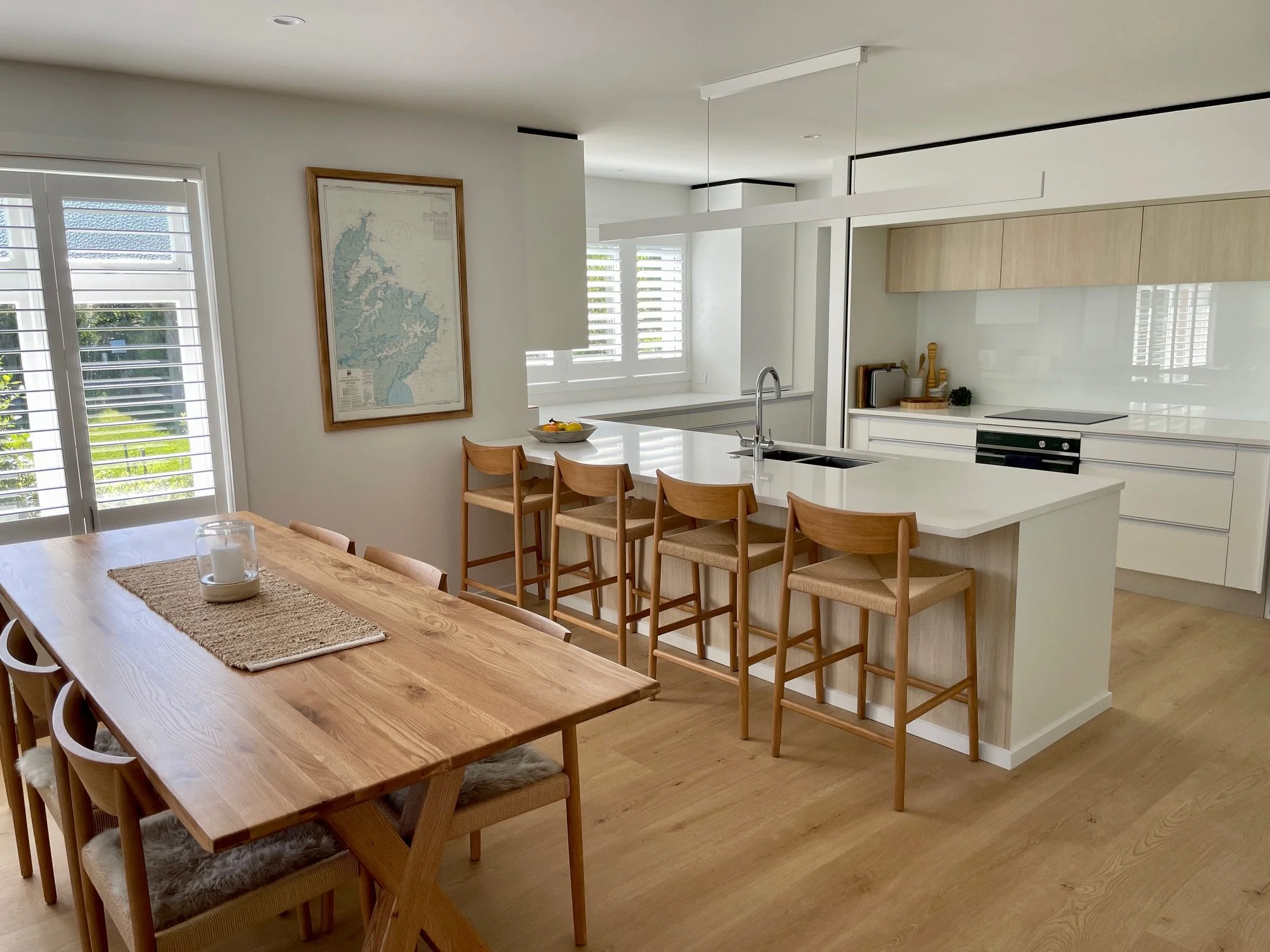 A modern open kitchen with white cabinetry, a large island with seating, and a farmhouse dining table with chairs. There are white shutters on the windows, and a framed map hangs on the wall.