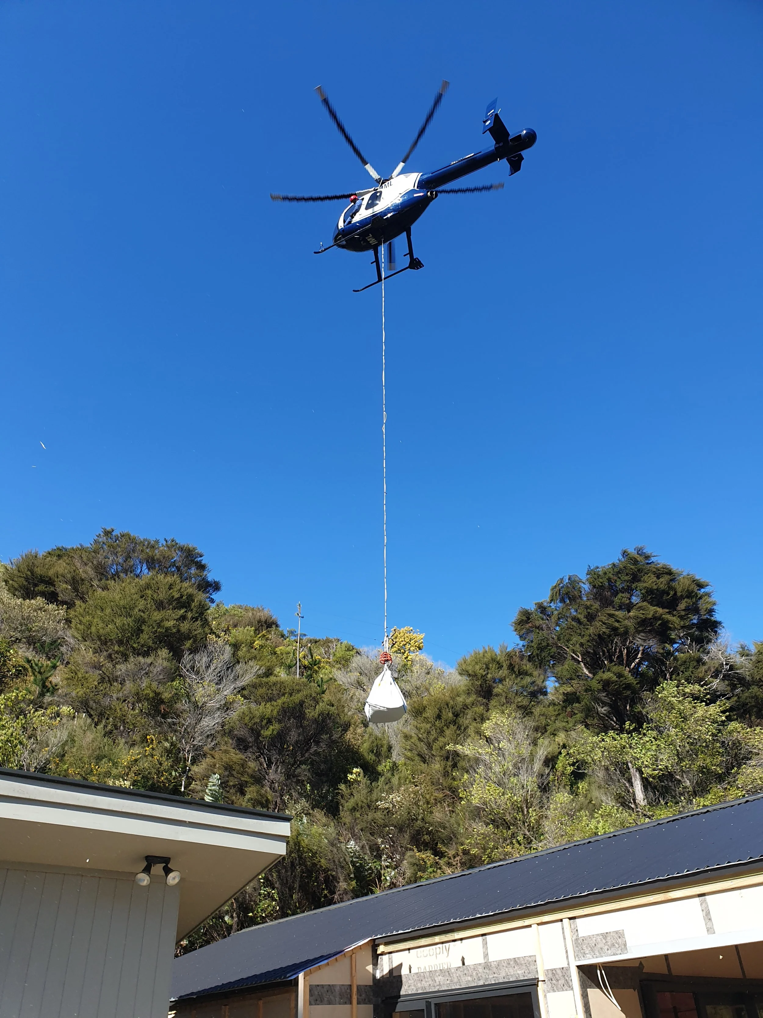 A helicopter hovering in the sky with a bucket hanging beneath it, possibly for firefighting or utility purposes. There are buildings and trees in the foreground and a clear blue sky.
