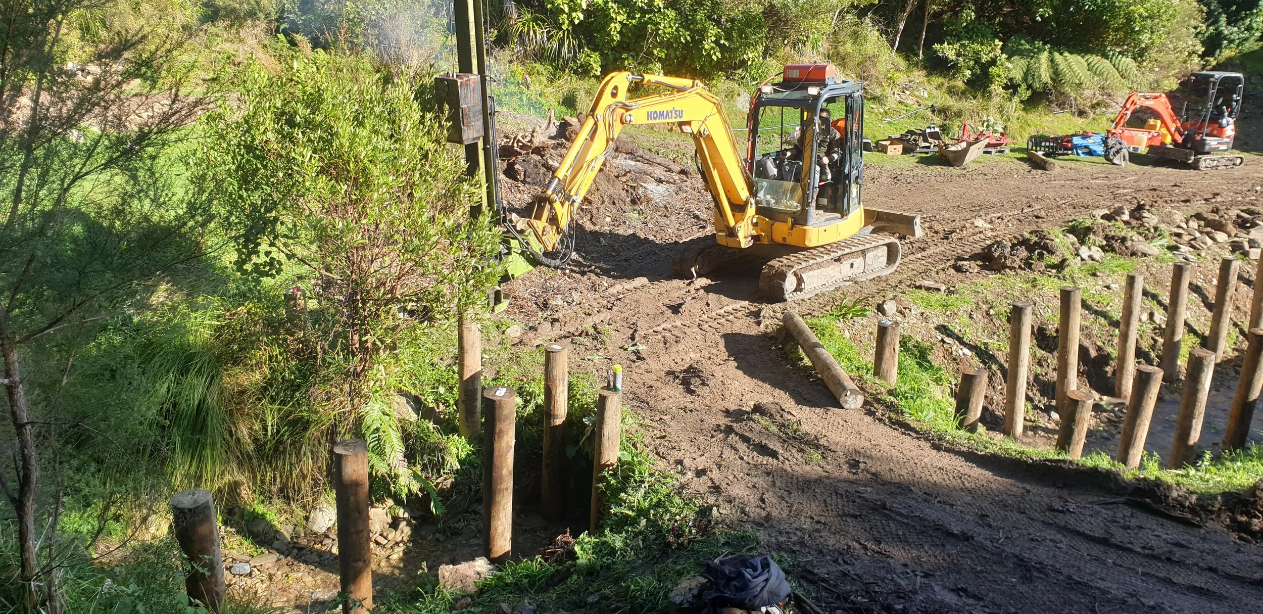 Construction site with a small yellow excavator digging into the ground, surrounded by construction tools and equipment, trees, and a partially muddy pathway.