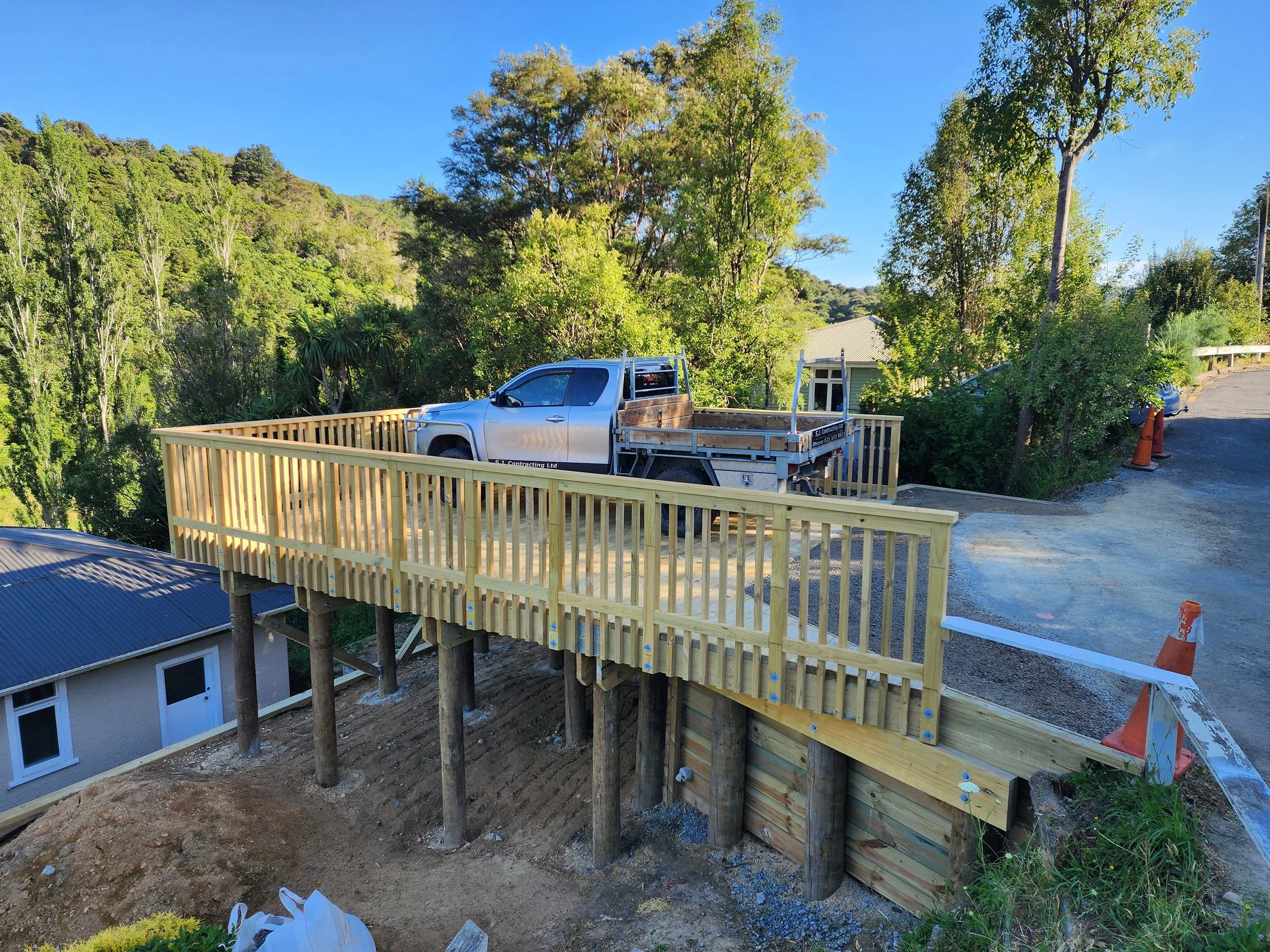A construction site showing a wooden deck with railings built on stilts, overlooking a hillside with trees. A pickup truck is parked on the deck, and there are construction cones and a ladder nearby.