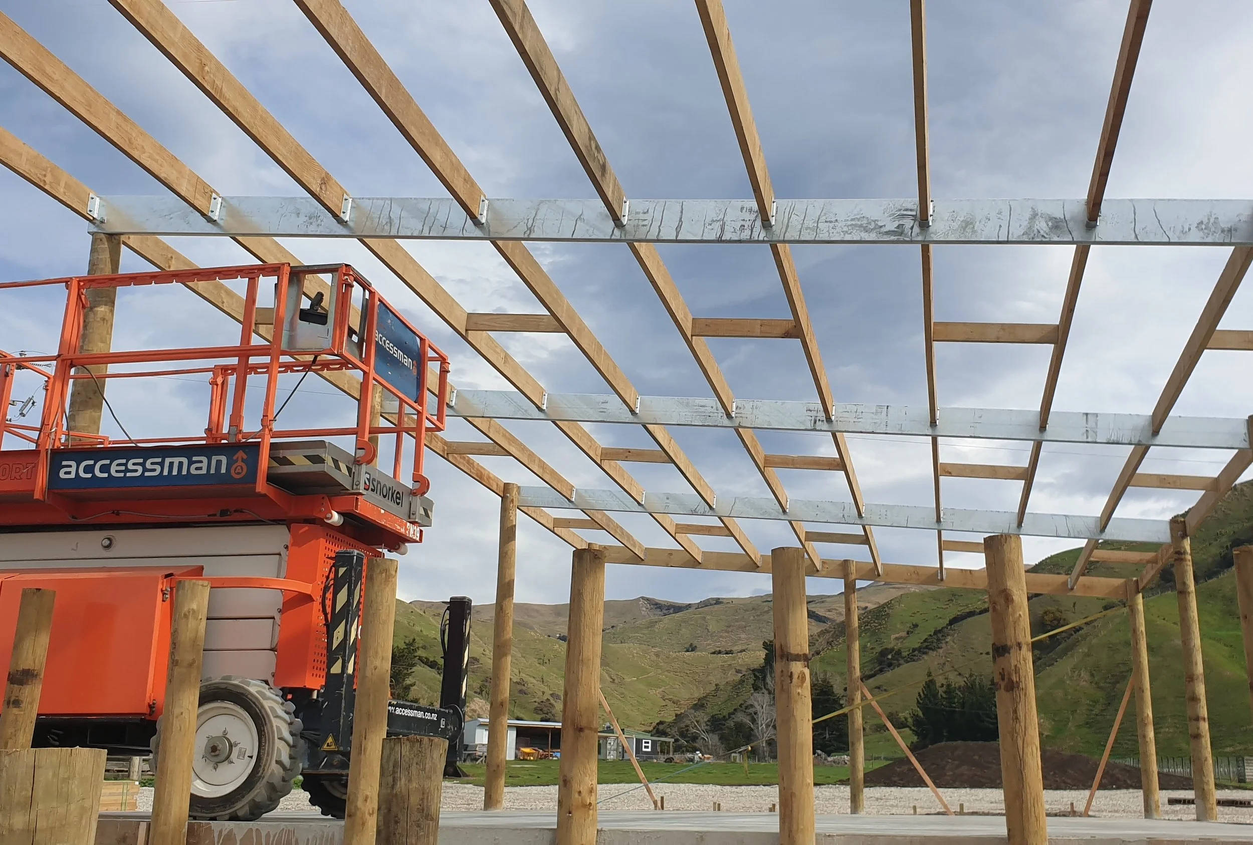 Construction site with wooden framing and a cherry picker lift in a rural area, with green hills and scattered houses in the background.