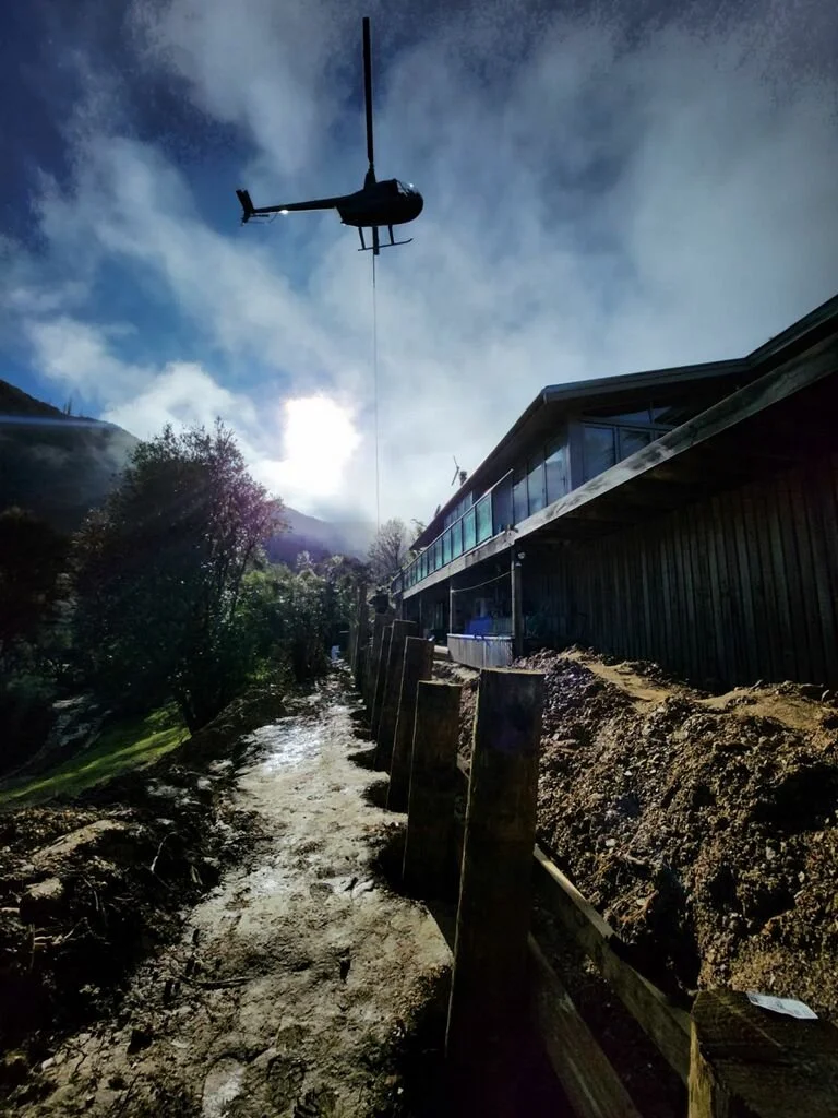 A helicopter flying in the sky above a construction site beside a house, with a dirt path and trees in the background.