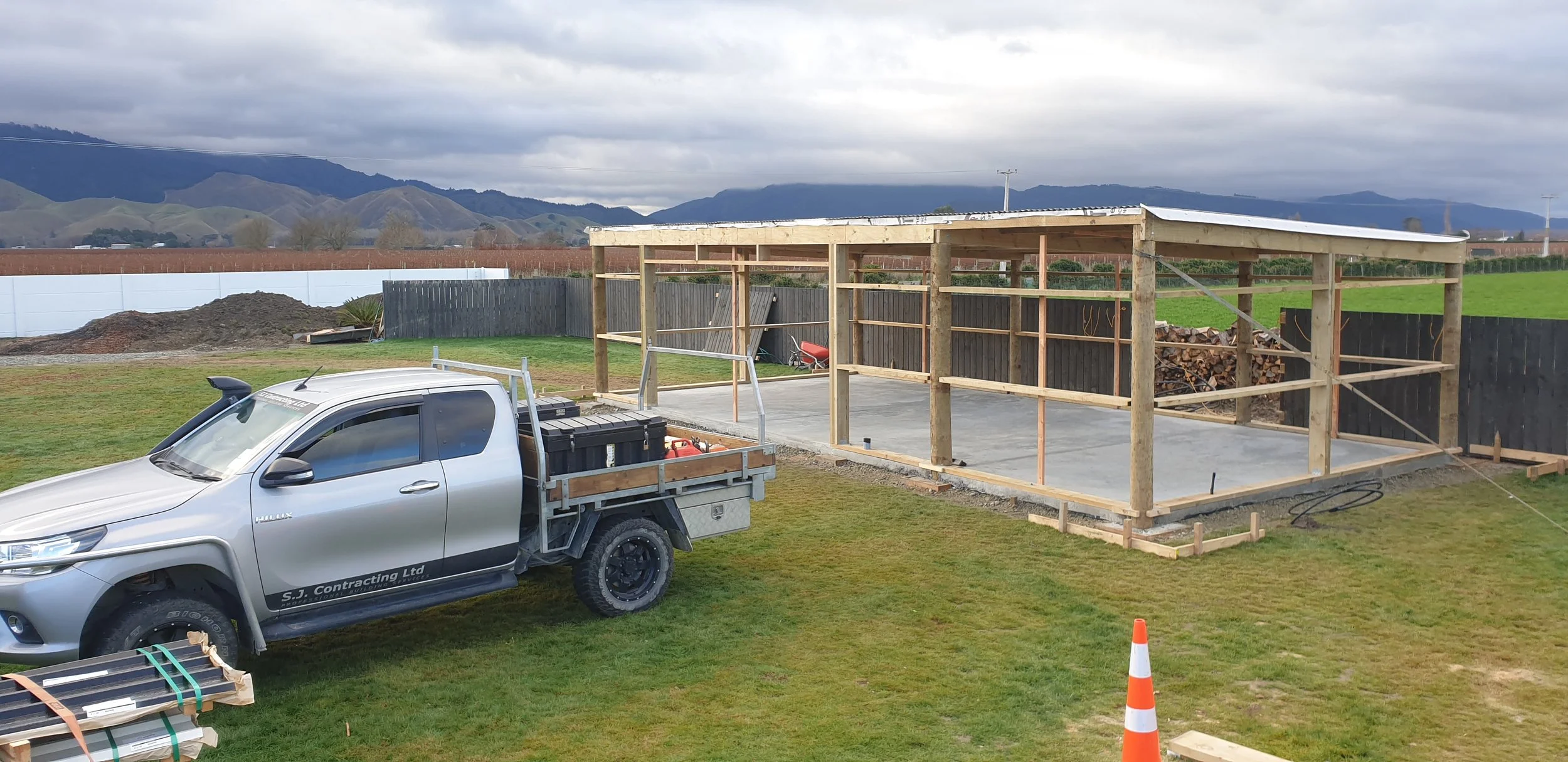 A construction site with a partially built structure on a concrete slab, wooden framing, a gray pickup truck with a flatbed, construction tools and materials nearby, and a scenic background of mountains and cloudy sky