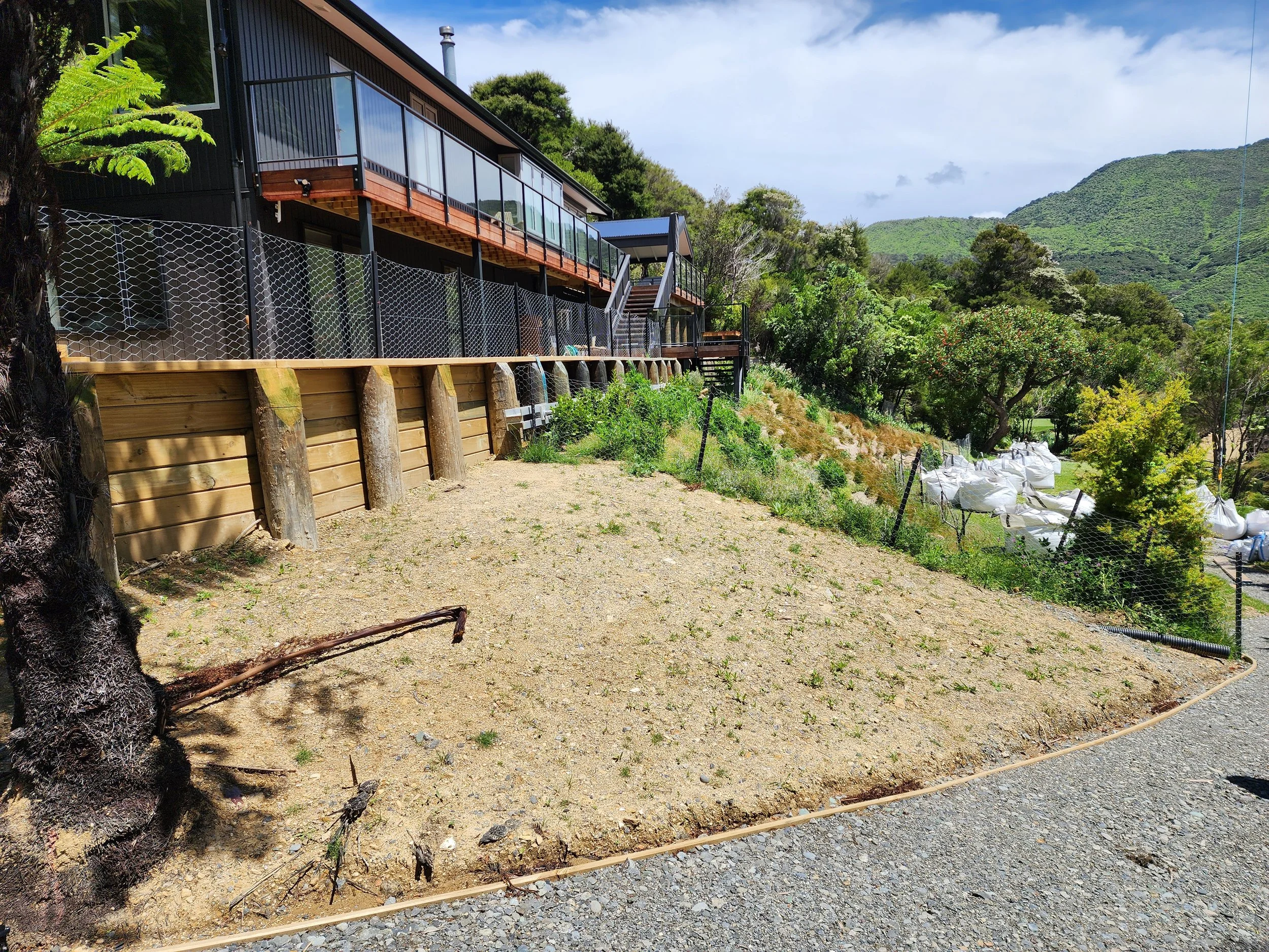 A modern black house with large glass windows sitting on a hillside surrounded by trees and mountains. There is a balcony with glass railing on the upper floor and a stairs leading to the yard. The yard is bare with some patches of grass and a gravel