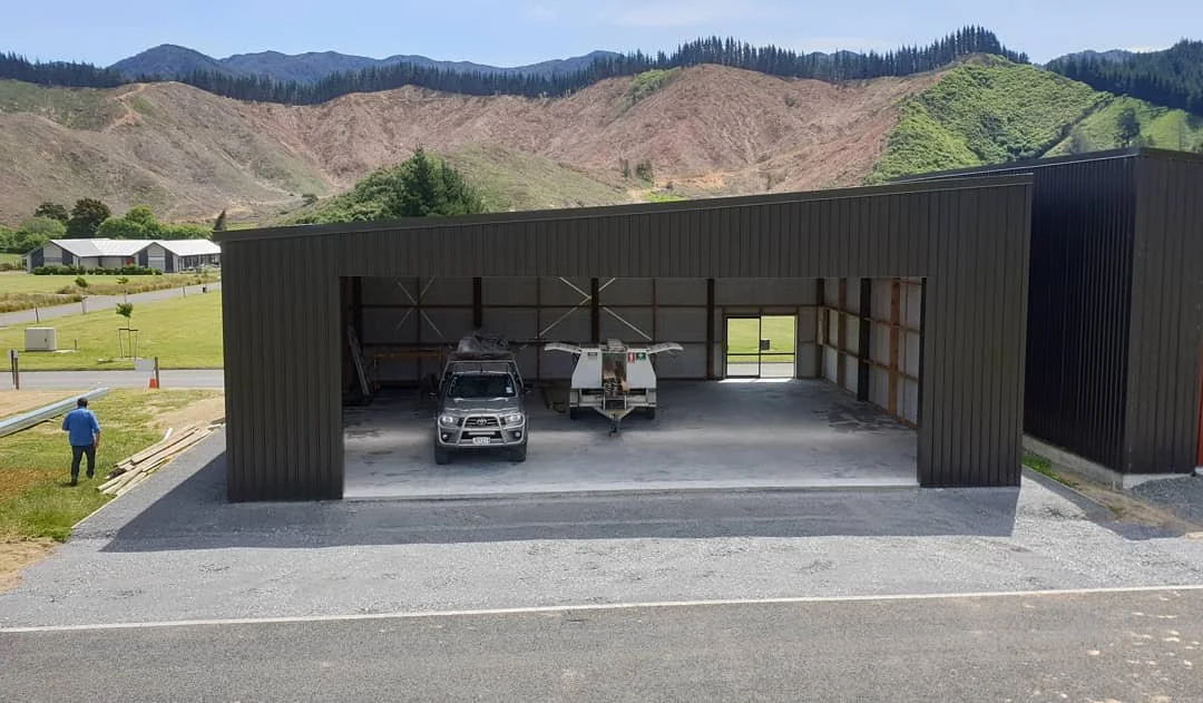 A partially constructed building with a black metal exterior, containing a parked car, a small plane, and building materials inside. A person is walking nearby, with mountains in the background.