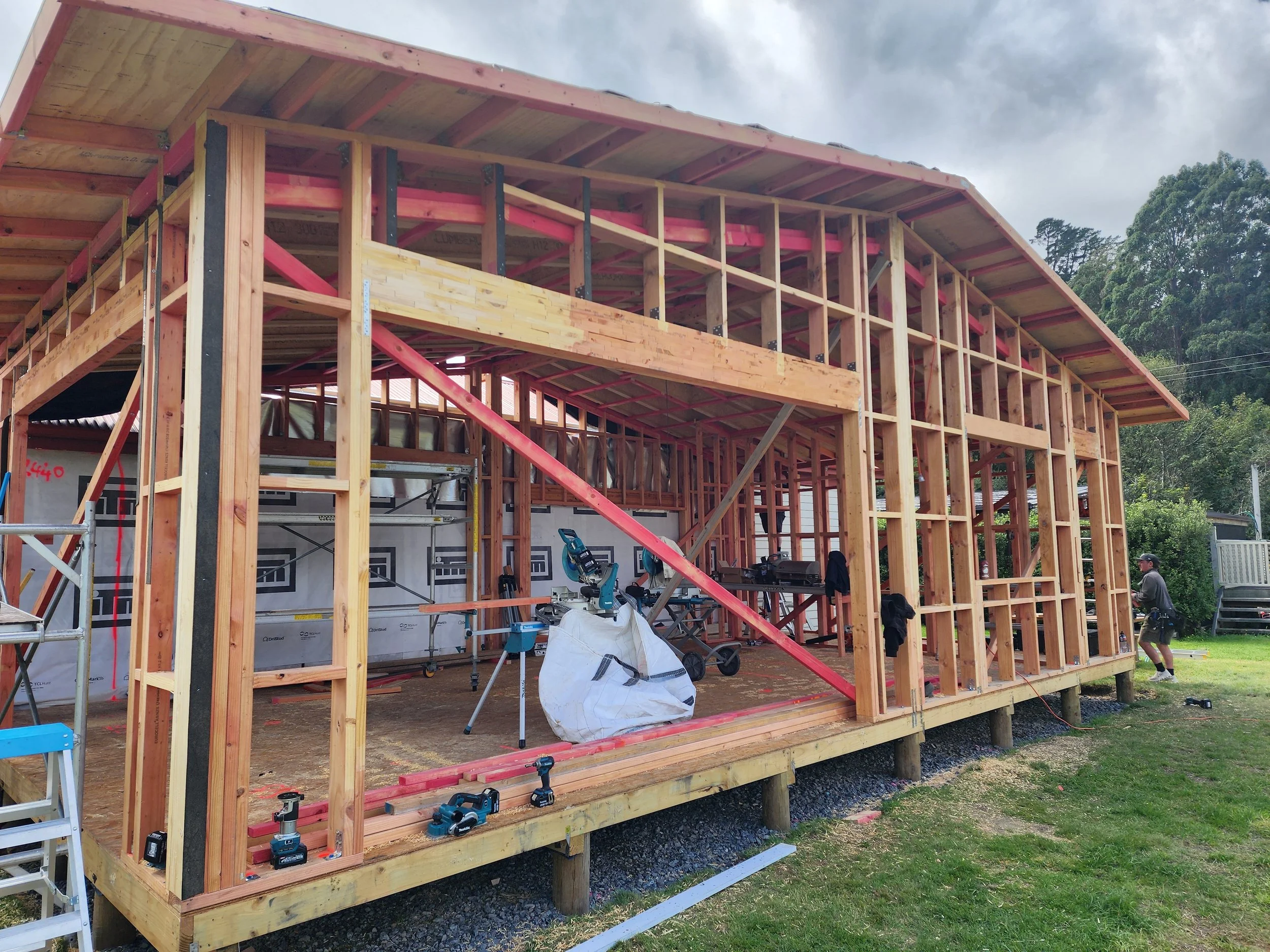 Wooden house frame under construction with tools and construction materials around, and trees in the background.