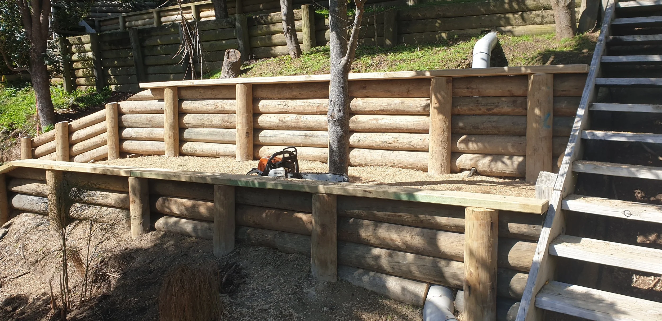 Wooden construction with stairs leading up to a raised platform, surrounded by trees and greenery, with a chainsaw placed on the ground.
