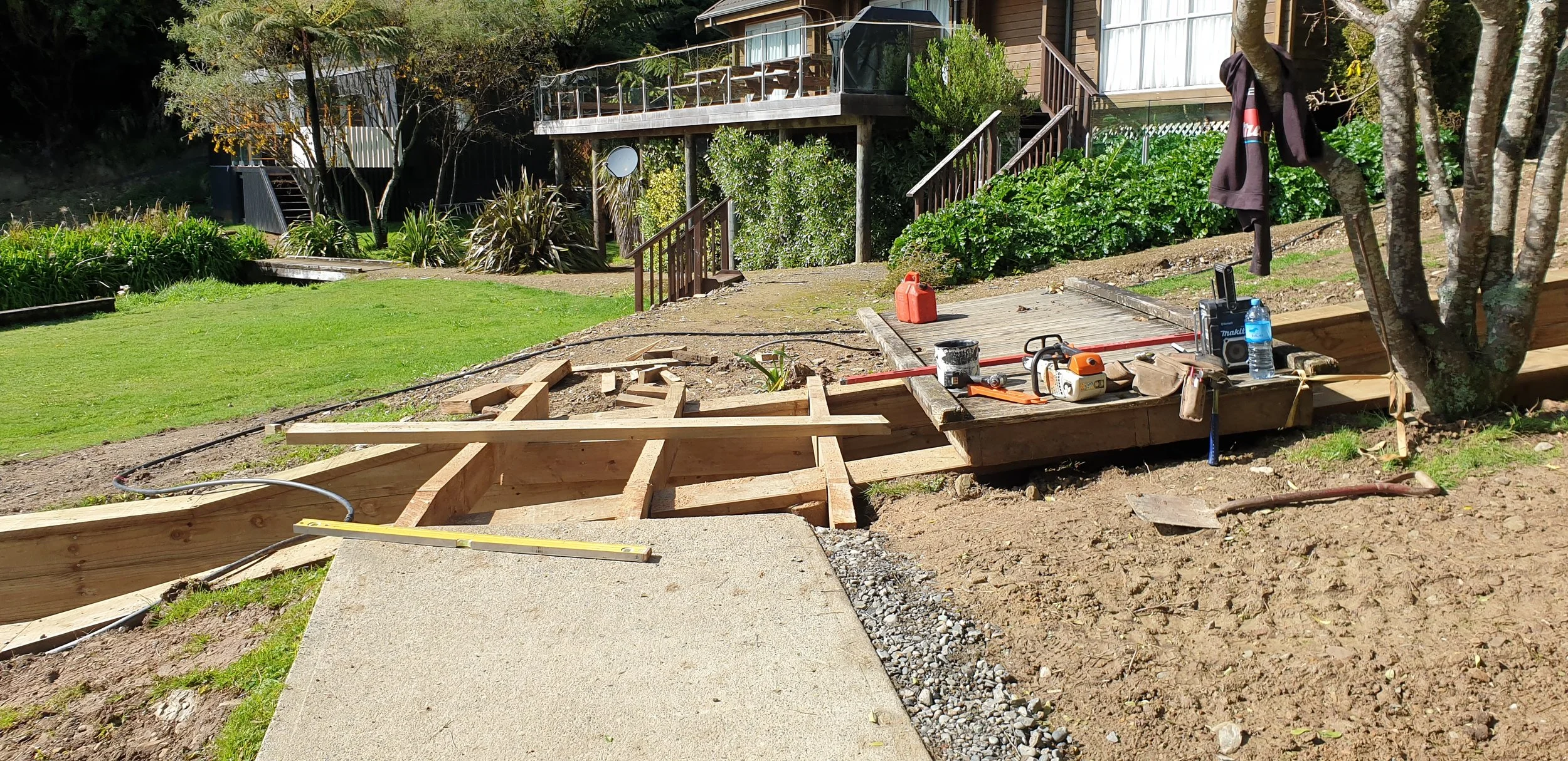 The image shows a backyard construction site where a wooden pathway or deck is being built. There are construction tools and materials, including a saw, level, and water bottle on the wooden frame. The background features a grassy yard, some trees, a