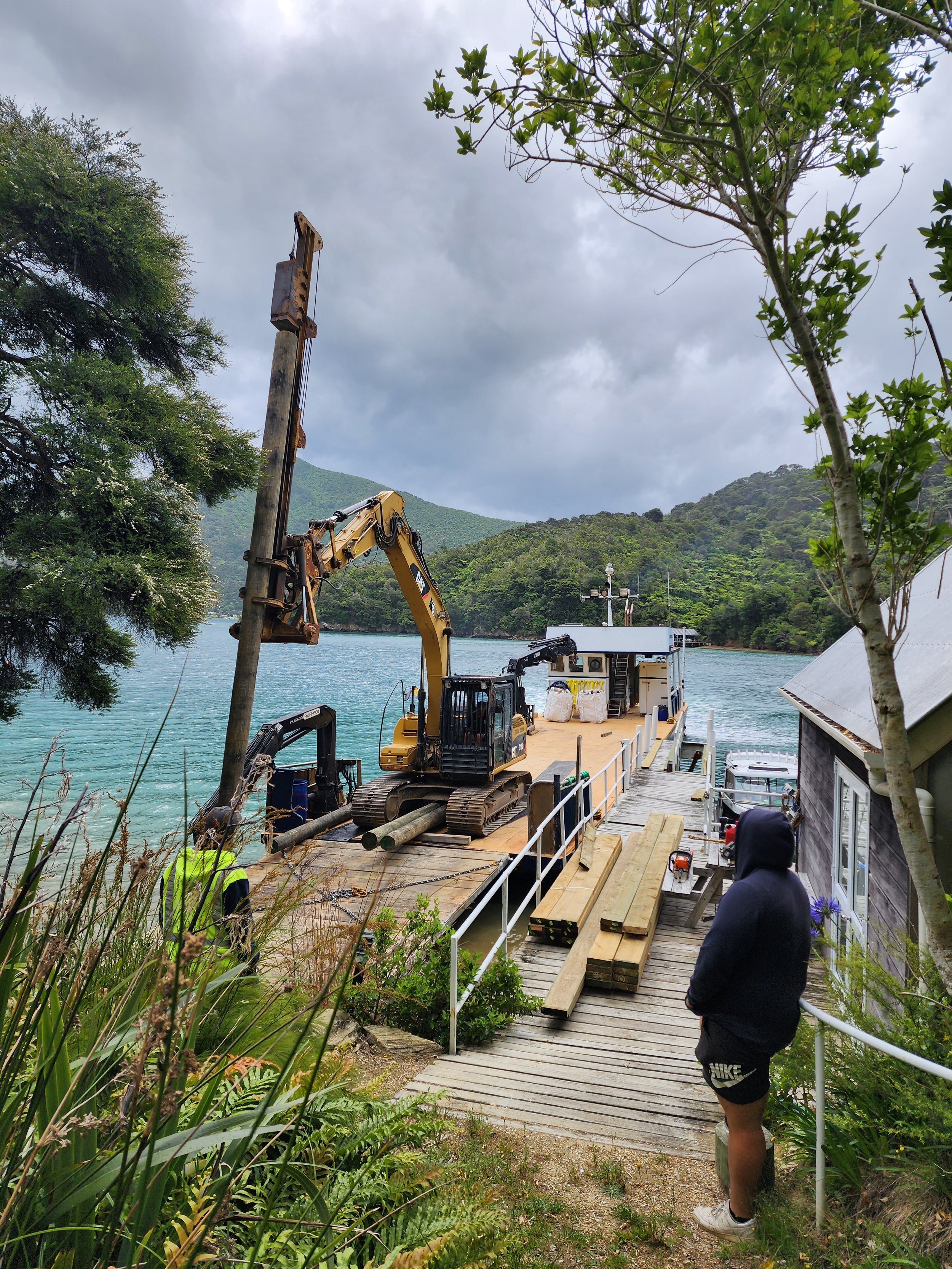 A construction worker and a person in hoodie near a dock with a small excavator, wooden planks, and equipment by a body of water surrounded by green mountains and trees.