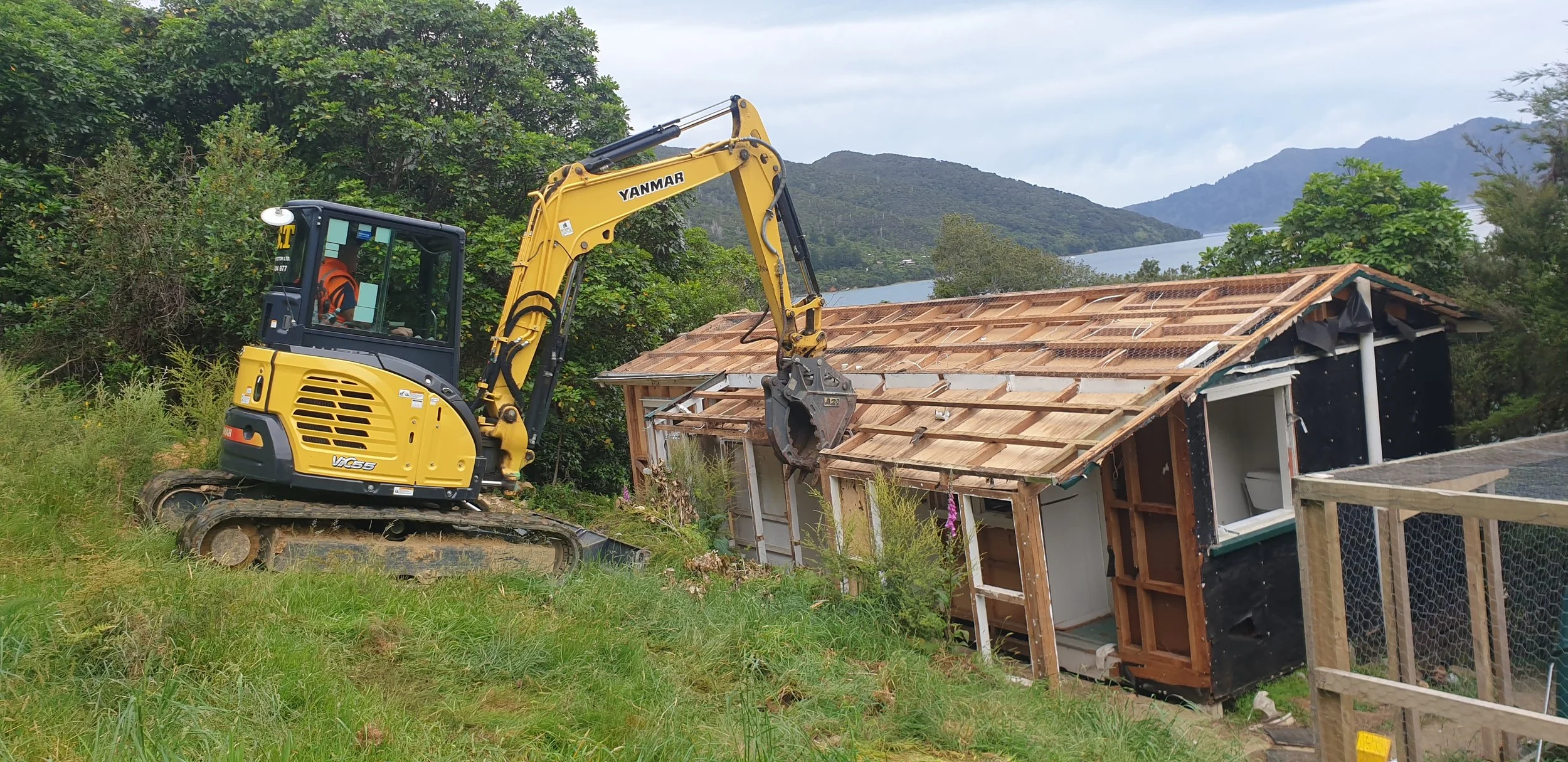 A yellow Yanmar compact excavator demolishing a small house with a wooden frame, black exterior, and a pitched roof in a green, mountainous landscape.