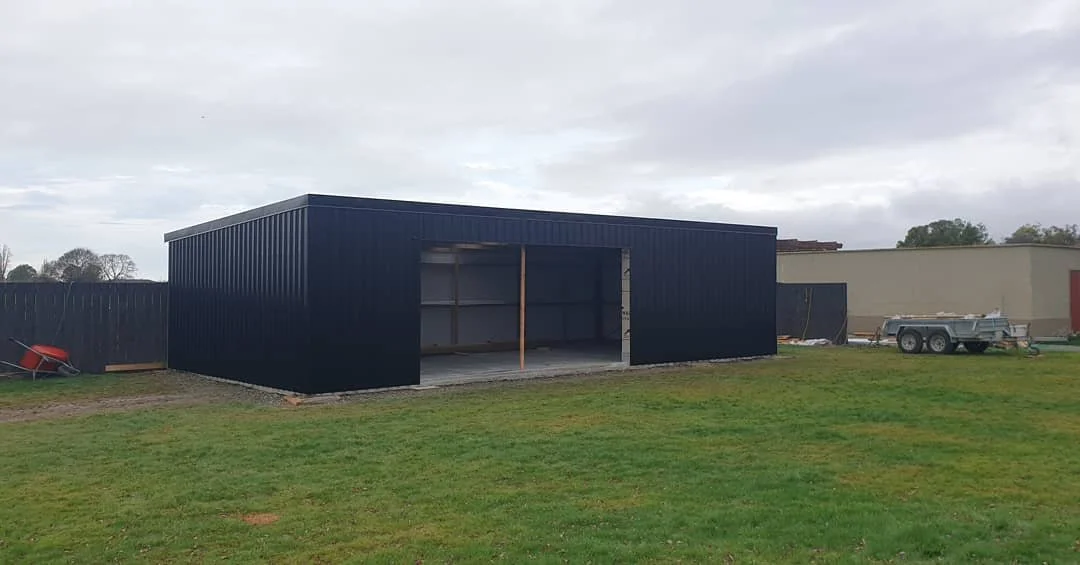 A small black metal shed under construction on a grassy area, with a wheelbarrow and trailer nearby.
