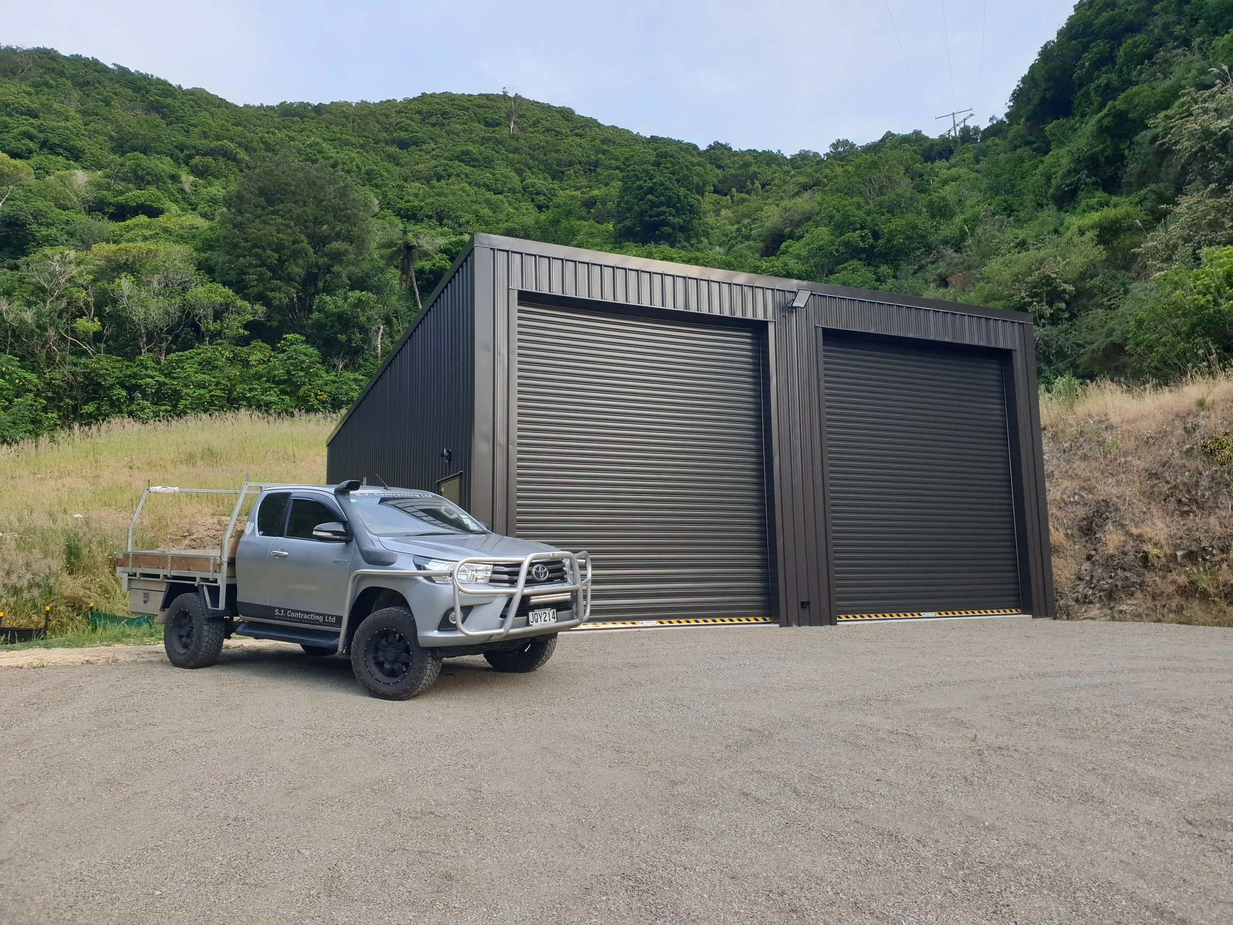 A gray pickup truck parked on the side of a road in front of a black industrial building with a rolling door, with a lush green hillside and trees in the background.