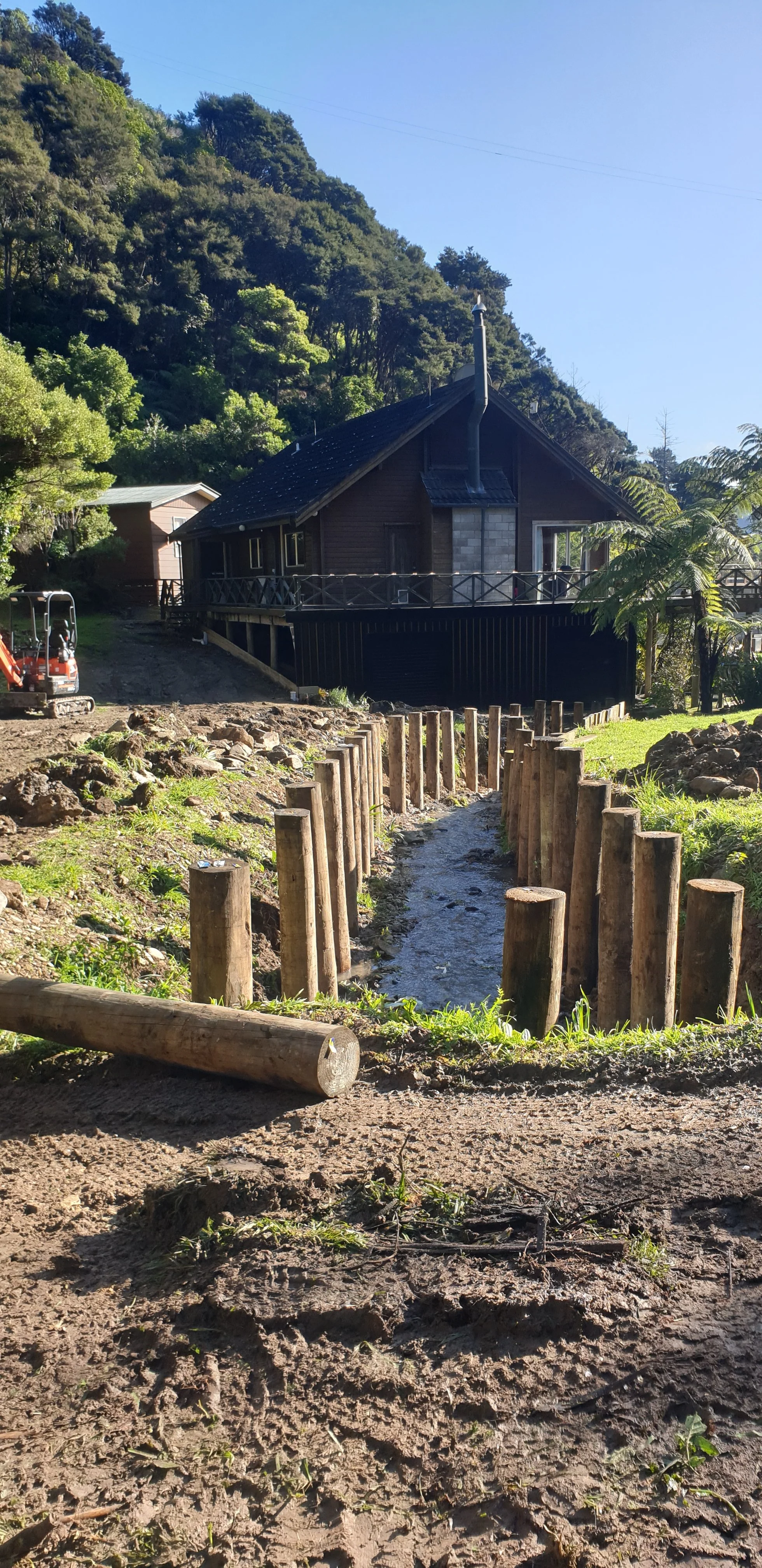 A small stream of water flowing through a muddy area with wooden posts lining the sides, in front of a dark wooden house on a hillside surrounded by trees and lush greenery.