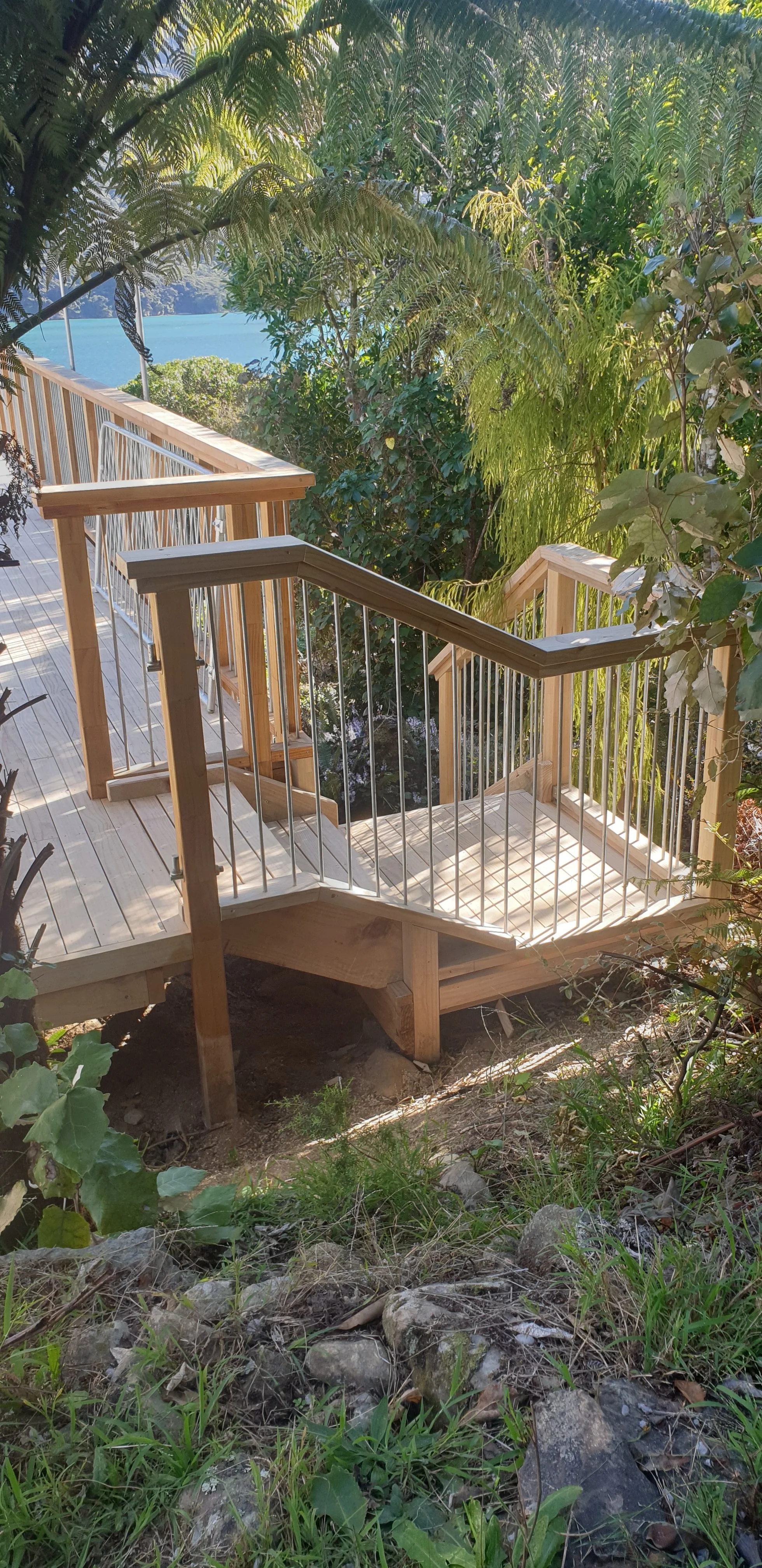 Wooden deck with metal railings surrounded by green foliage and trees, overlooking a body of water in the background.