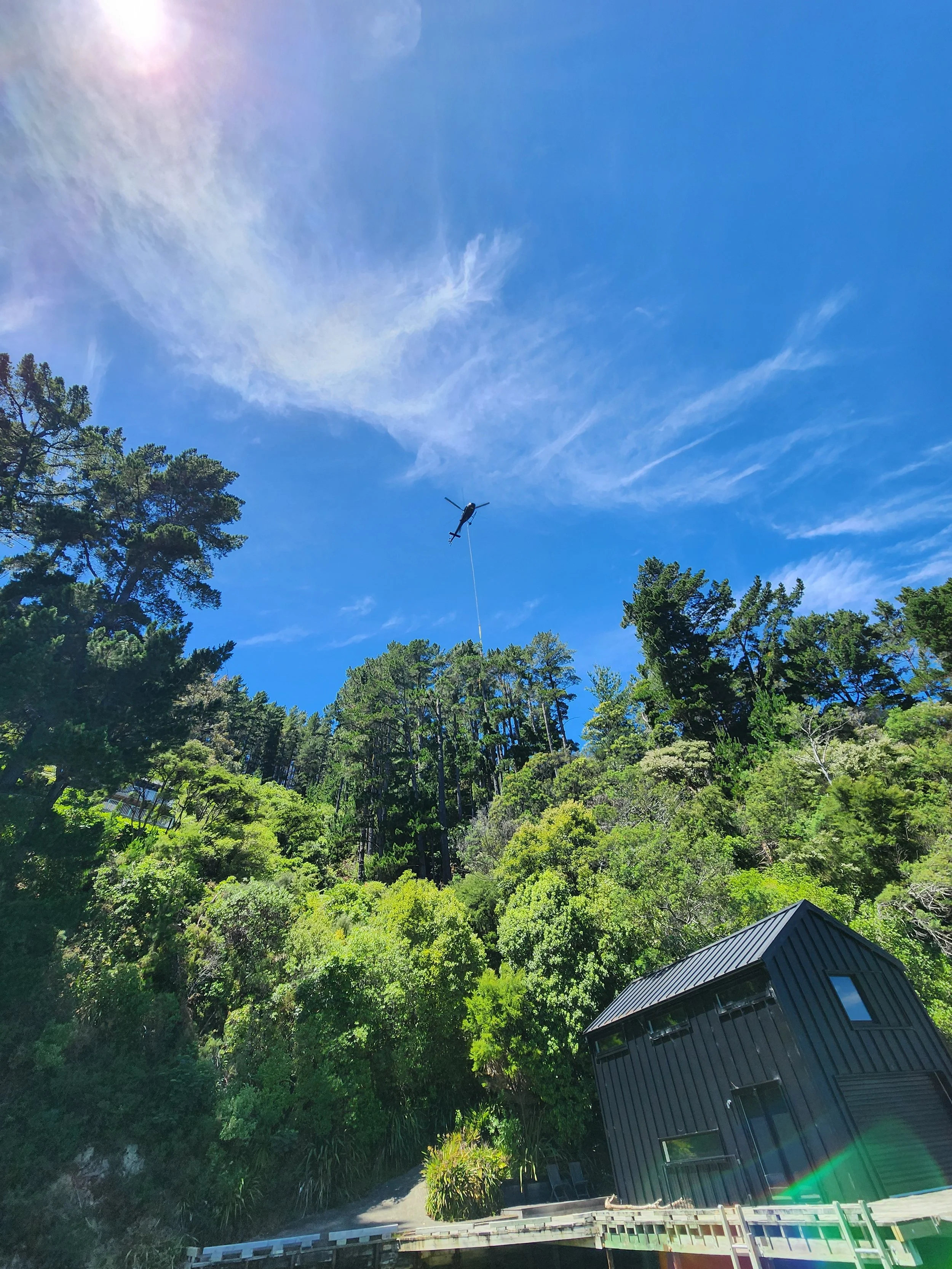 A helicopter flying in a clear blue sky above a lush green forest with a black modern building at the bottom right corner.