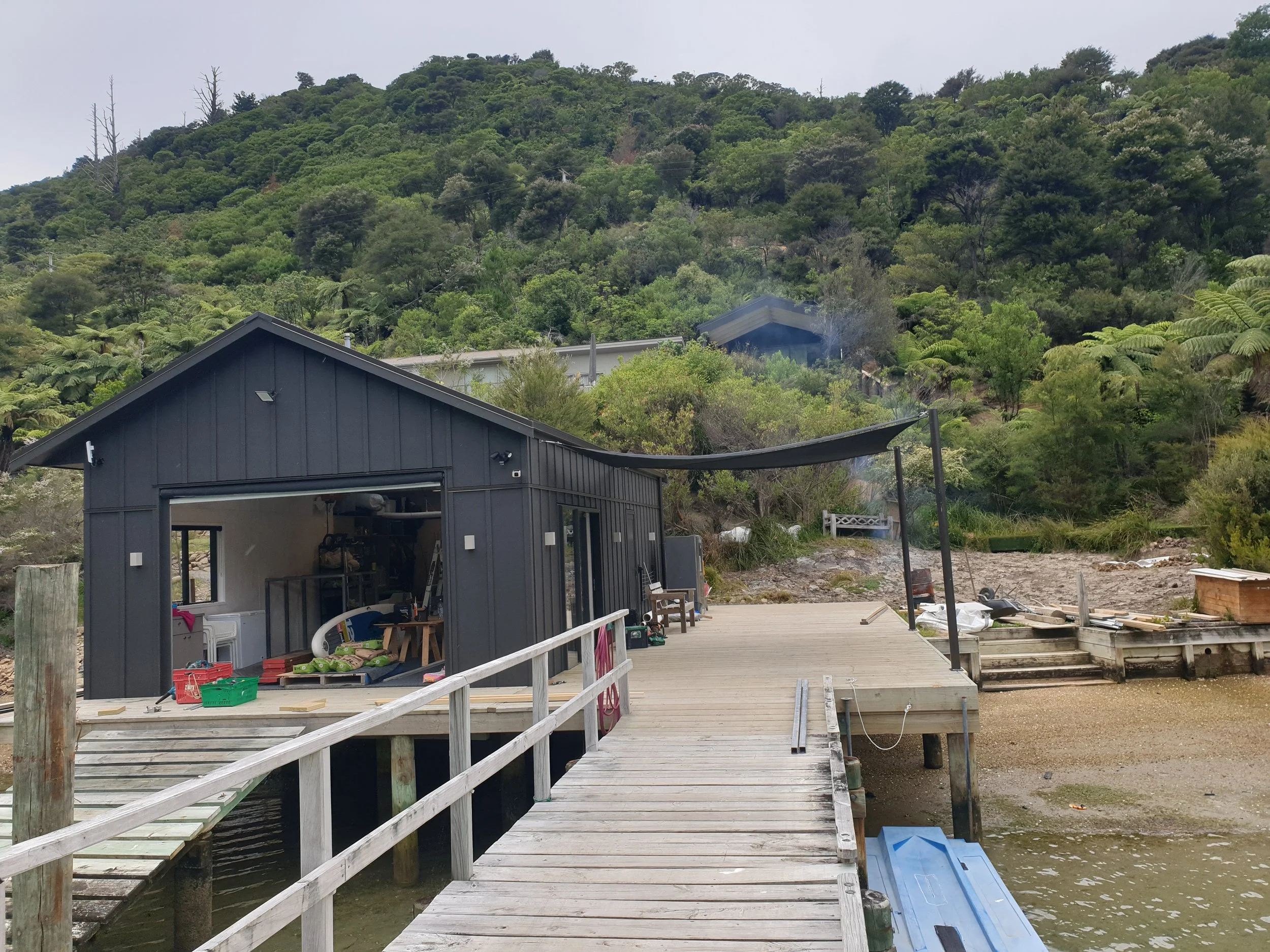 A house with a dark exterior situated near a marina, with a wooden dock extending into the water in the foreground. Behind the house, there is a lush green hillside with dense trees.