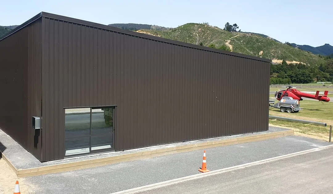 A modern dark brown metal building with a sliding glass door, set on a gravel and paved surface with an orange traffic cone nearby, surrounded by grassy fields, a red helicopter and a silver trailer in the background, with green hills and trees in th