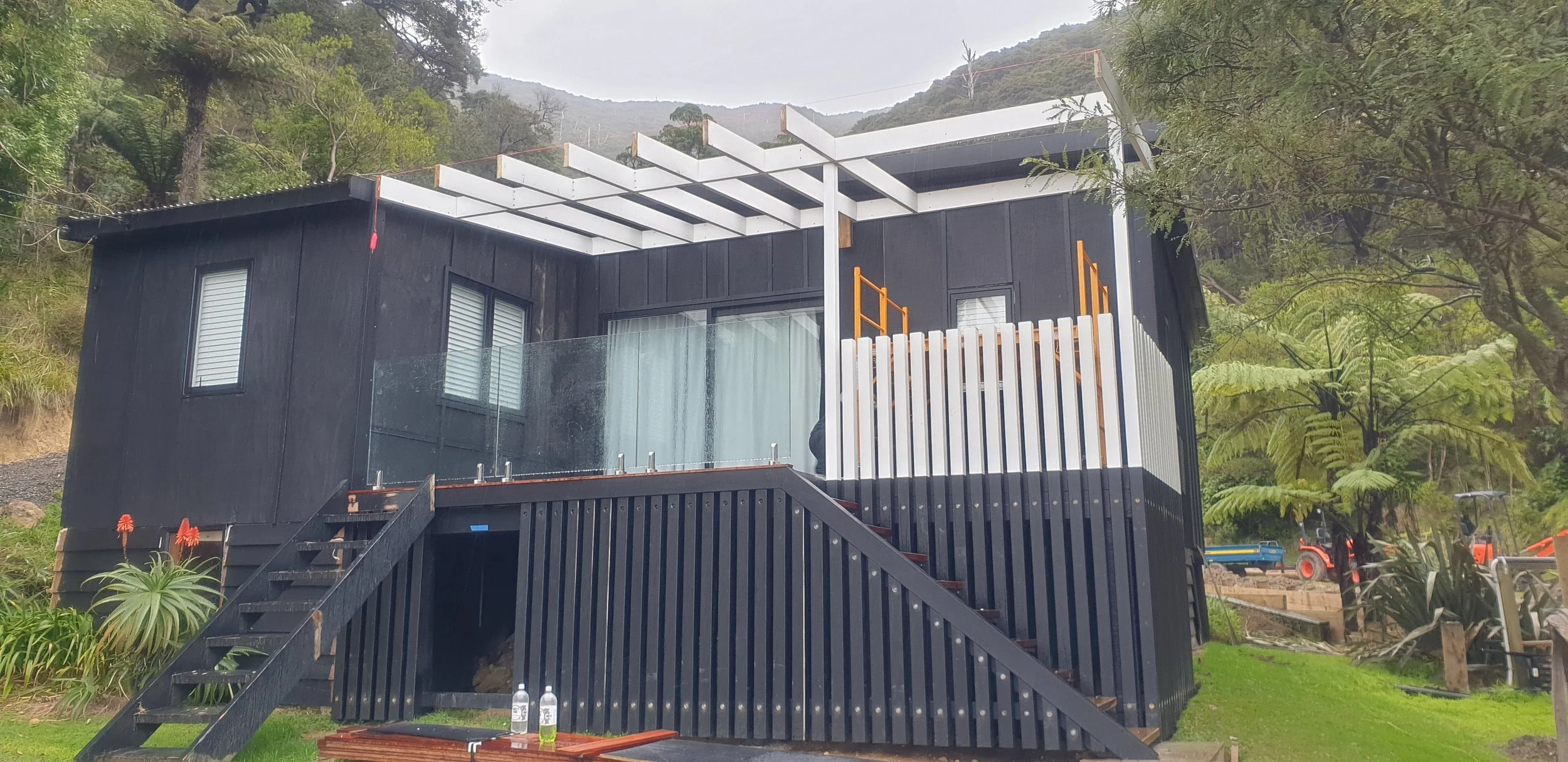 A modern two-story house with black exterior walls, large windows, and a glass balcony. The house is under construction with visible white beams on the roof. It is surrounded by trees and mountains, with construction equipment and tools nearby.
