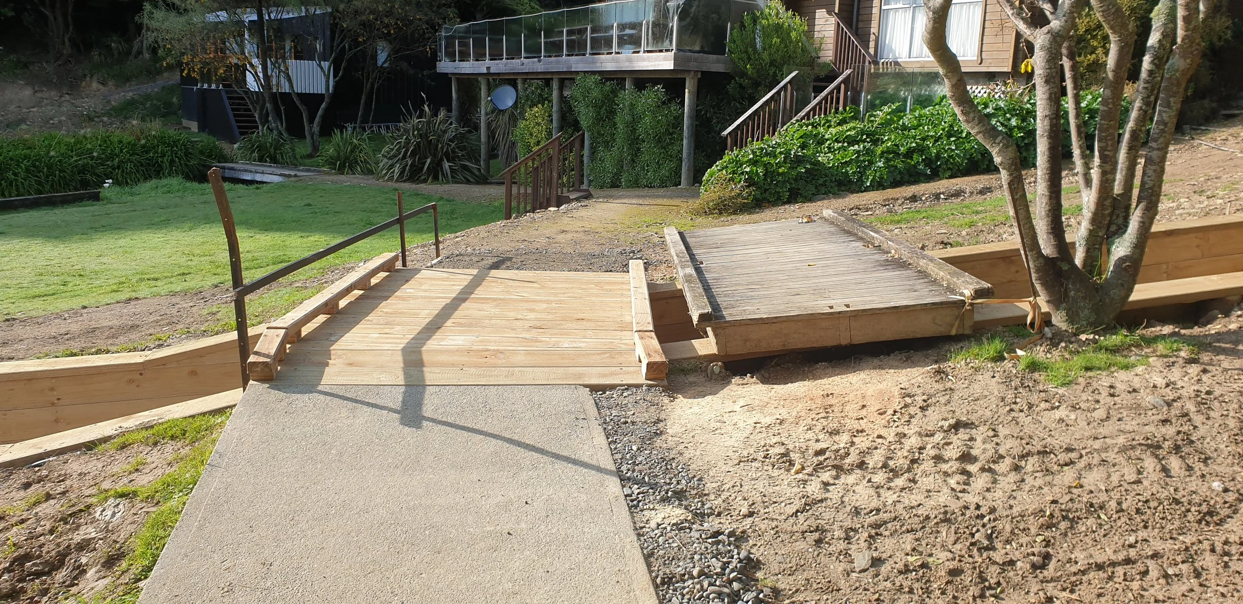 A newly built wooden wheelchair accessible ramp and sidewalk leading to a house, with green grass, trees, and shrubbery surrounding the area.