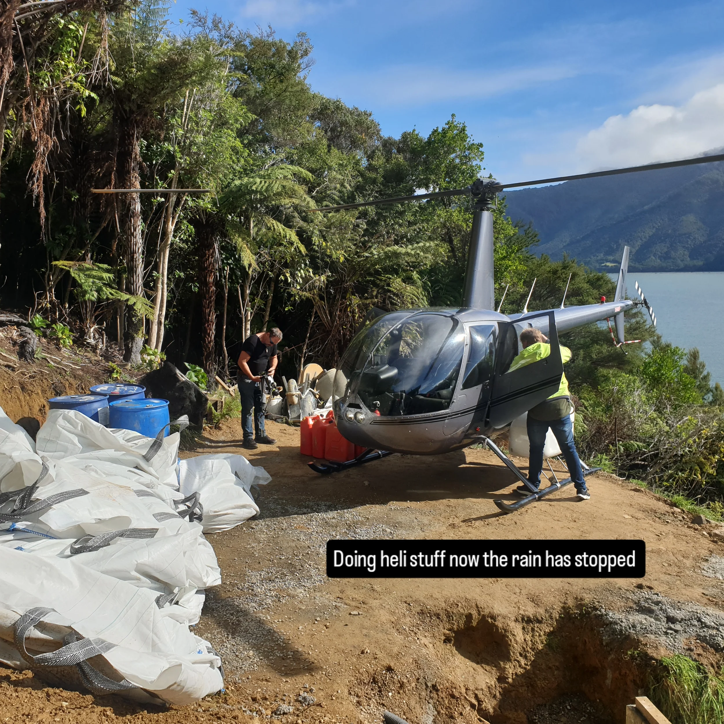 People working on helipad construction near a helicopter with a mountain and forest background. The scene shows clearing and ground preparation.