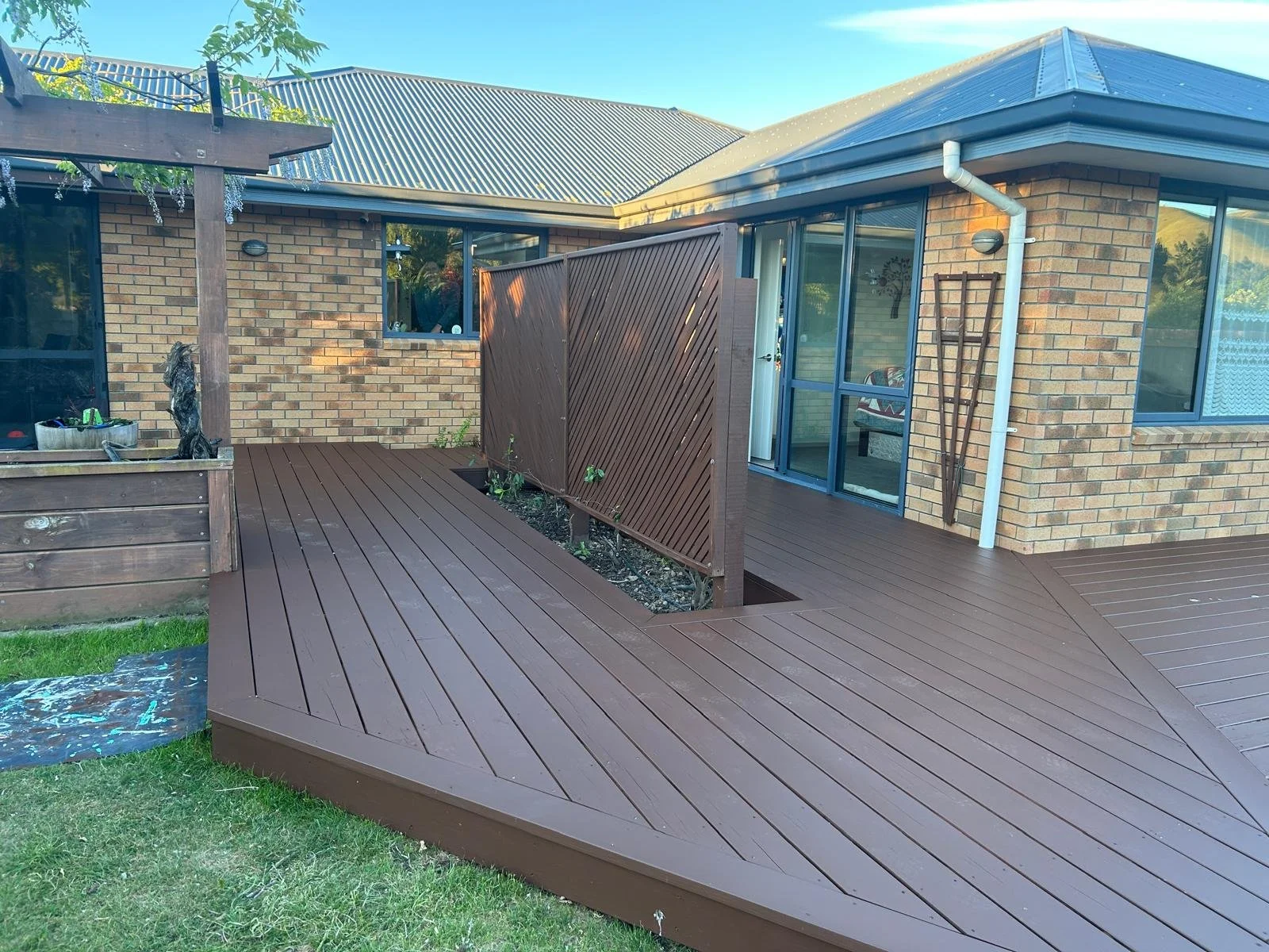 Backyard with a newly installed brown wooden deck, brick house with large windows, and a detached fencing section.