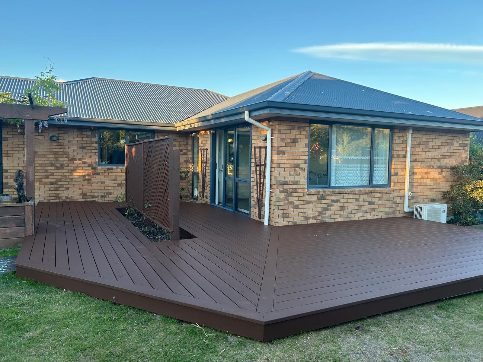 Backyard with a raised wooden deck attached to a brick house, featuring sliding glass doors and windows, with a small garden bed and a wooden privacy screen, under a clear blue sky.