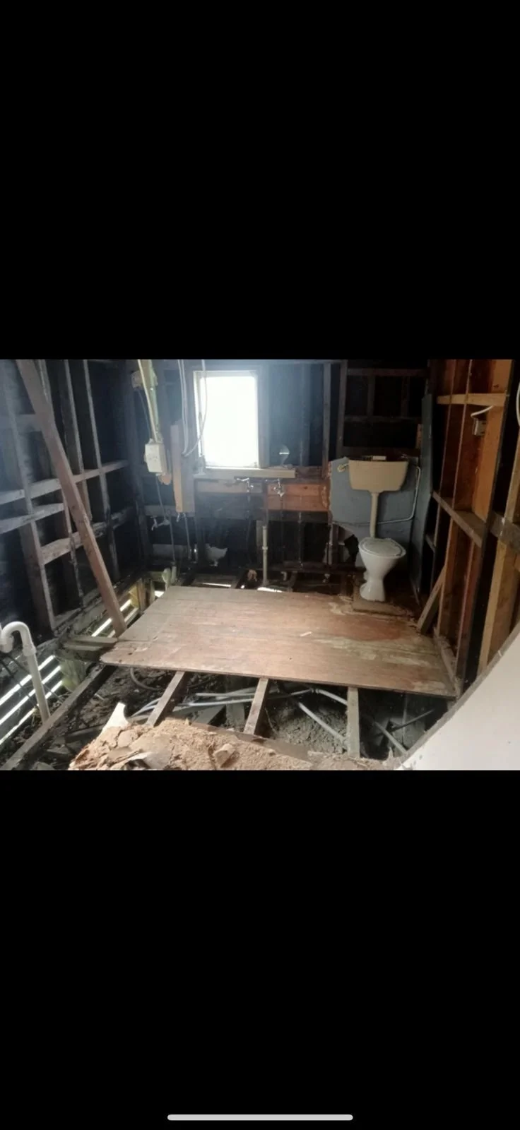 Bathroom under renovation with exposed wooden framing, a toilet, a small window, and plumbing. The floor is partially removed, revealing floor joists and construction debris.
