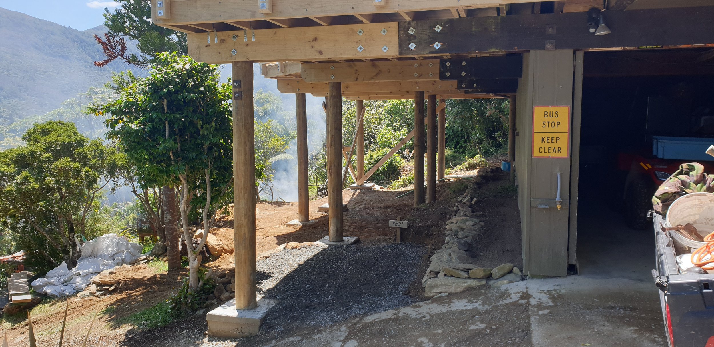 Construction site of a building with wooden support beams, a parking area, and a yellow sign that reads 'Bus Stop Keep Clear'. There's a dirt path and trees in the background.