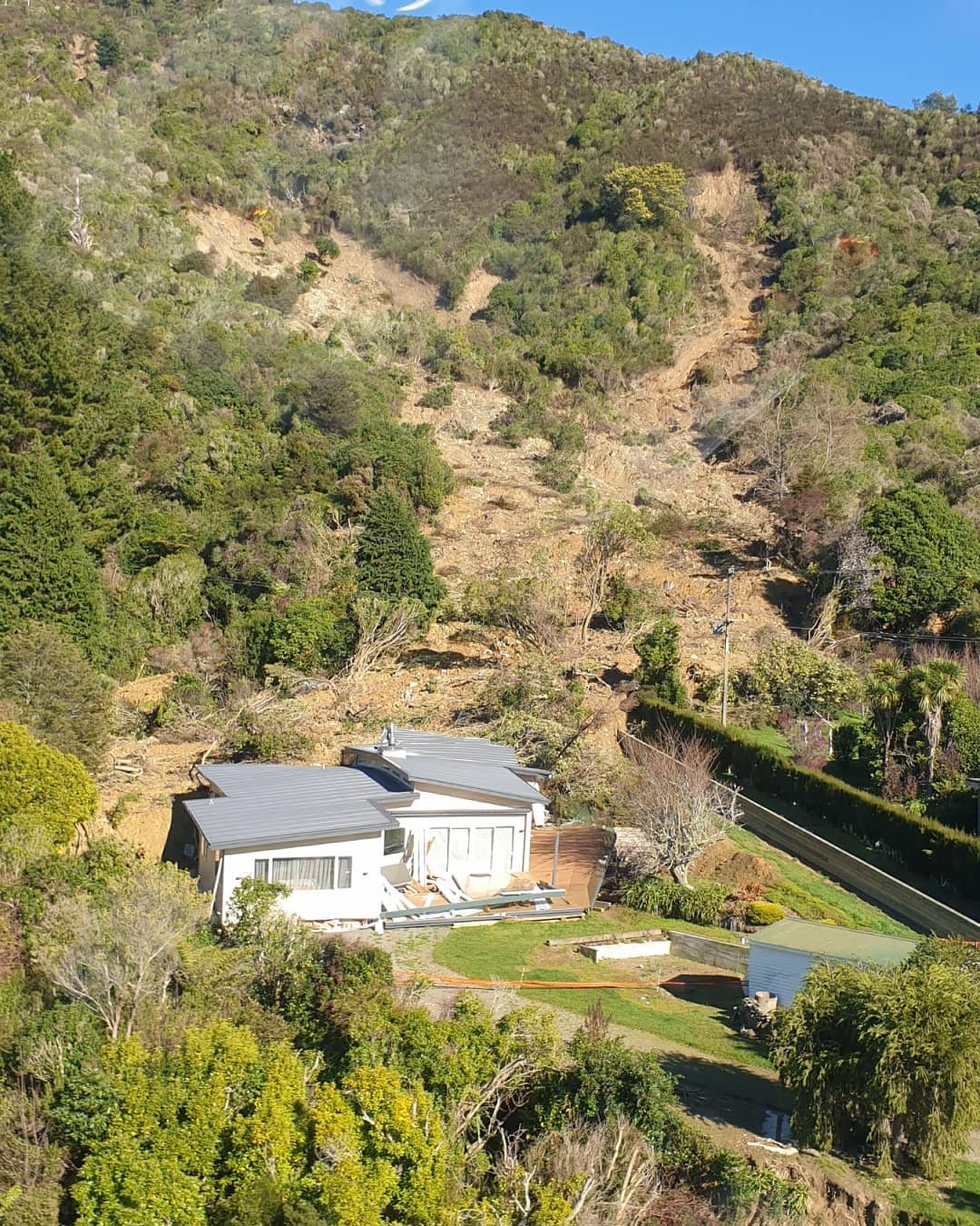 A house with a damaged roof and siding in a hilly area with green trees and a clear blue sky.